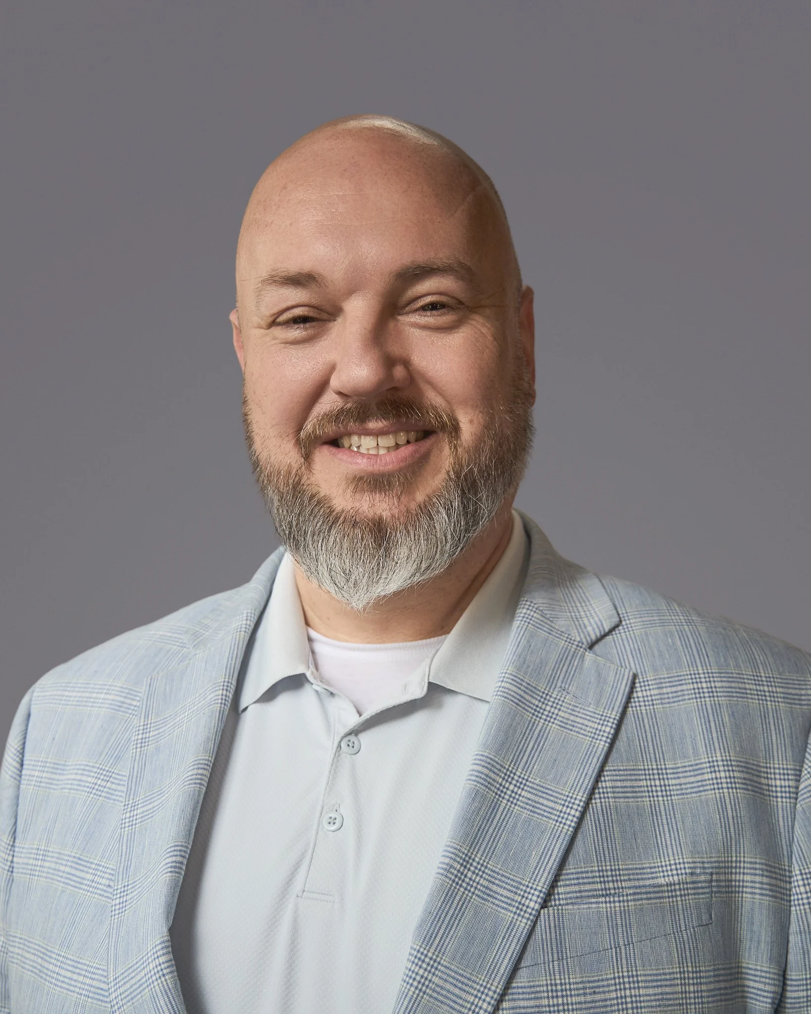 Close-up of a smiling man with a bald head, full beard, wearing a light blue plaid blazer, white shirt, and a light gray background.