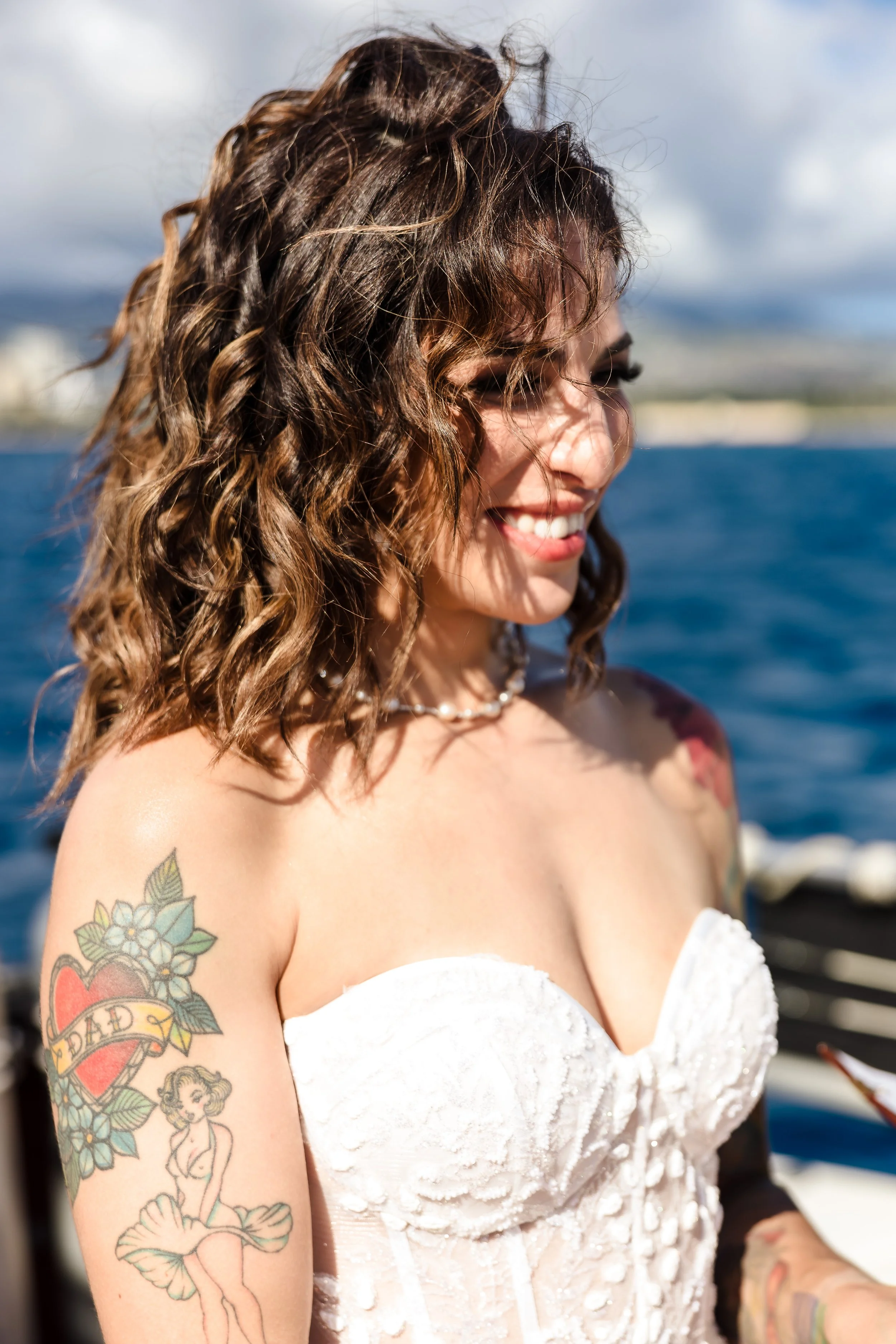 A woman with curly brown hair smiling outdoors, wearing a white strapless dress with a textured pattern, a pearl necklace, and tattoos on her upper arm, against a backdrop of water and cloudy sky.