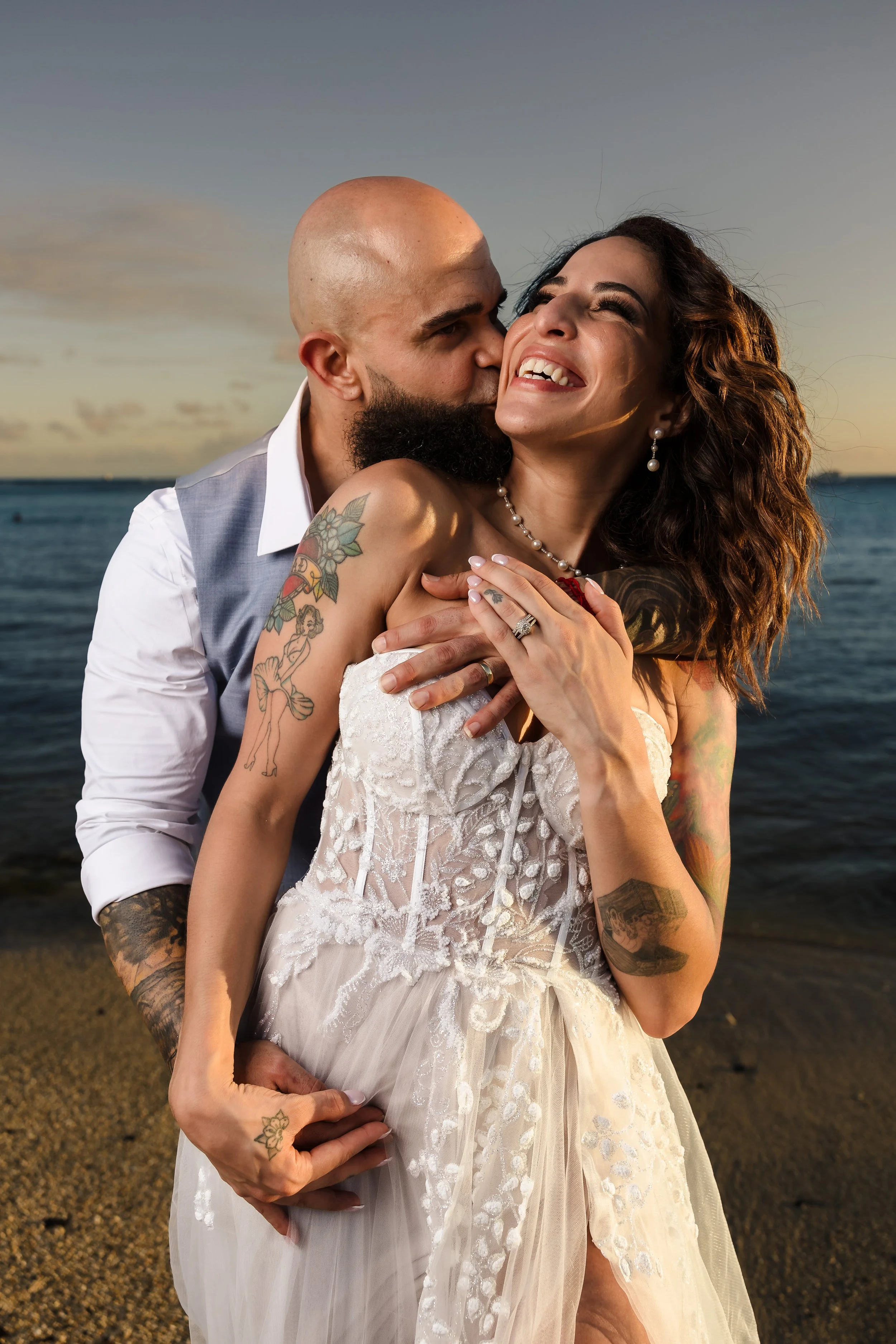 A happy couple dressed in wedding attire embracing by the beach at sunset, with ocean and sky in the background.