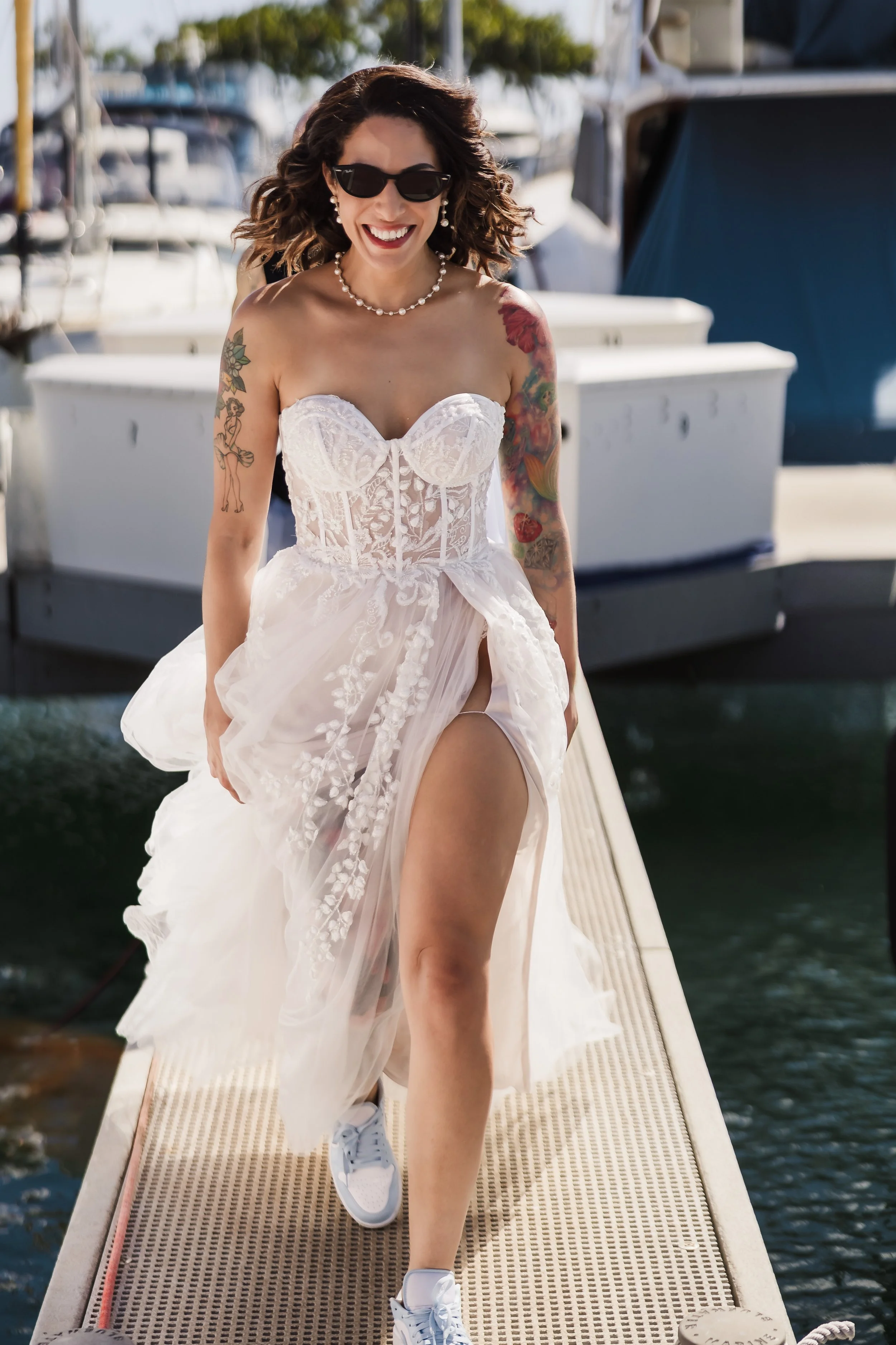 Woman in a white dress with tattoos, sunglasses, and pearls, walking on a dock near boats