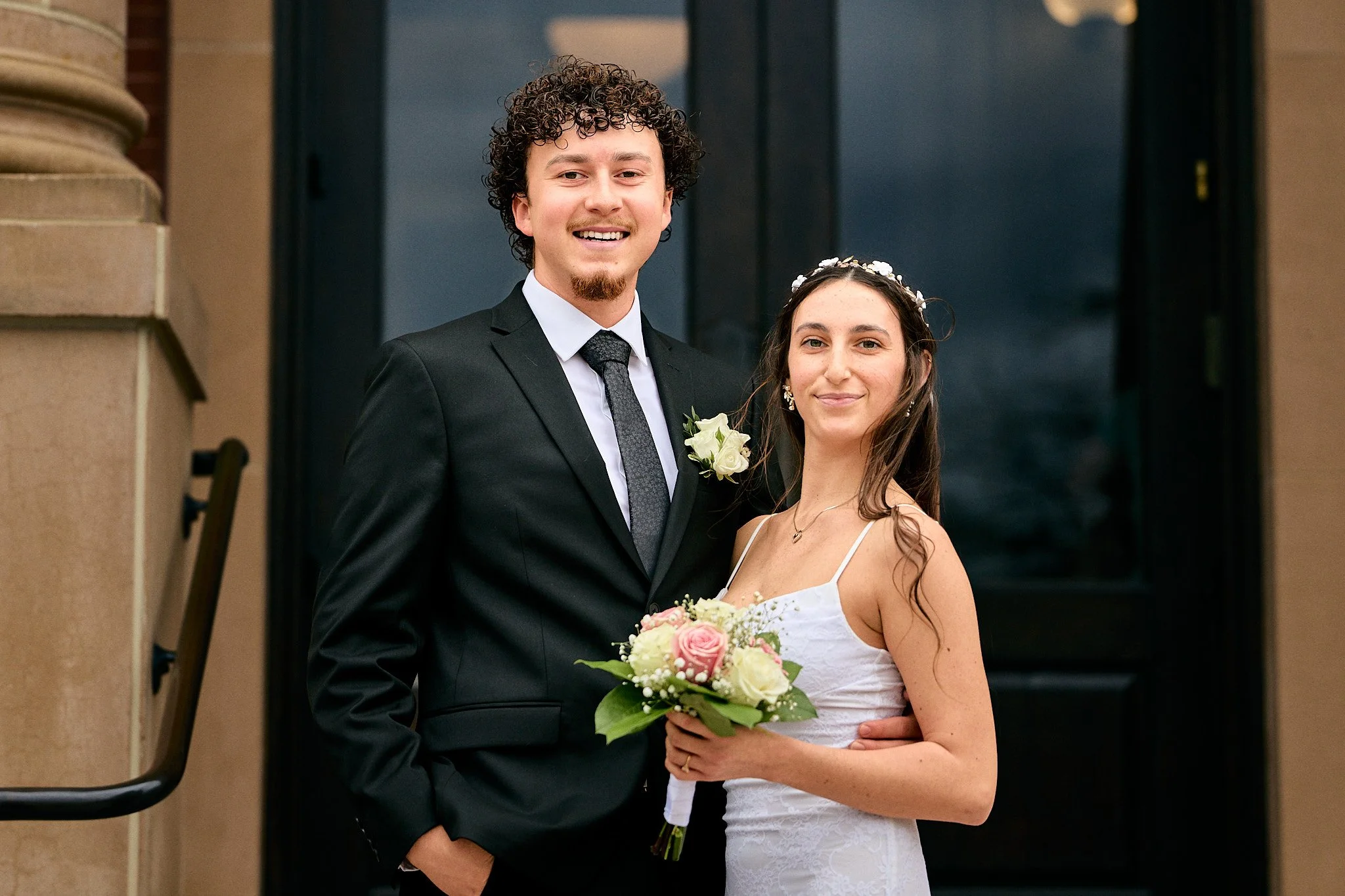 A newlywed couple stands together outdoors in front of a dark door. The groom has curly hair, a beard, and is dressed in a black suit with a white shirt and gray tie. The bride has long dark hair, wears a lace white wedding dress, and a floral headba