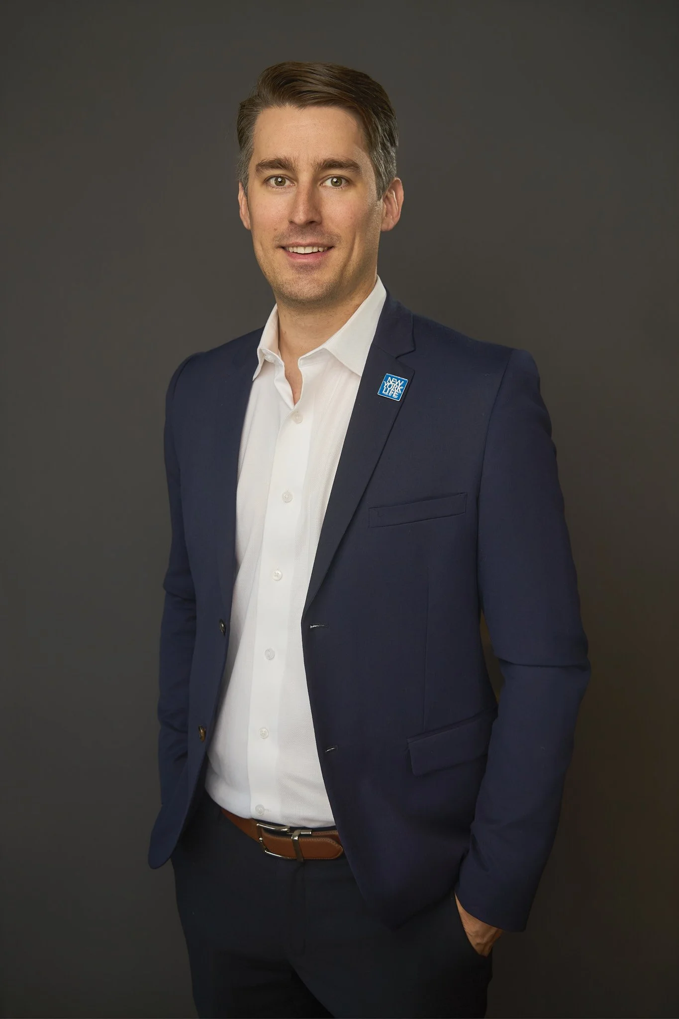 A man in a navy blue suit with a white shirt, smiling, with a lapel pin that says "New York Life" on a dark background.