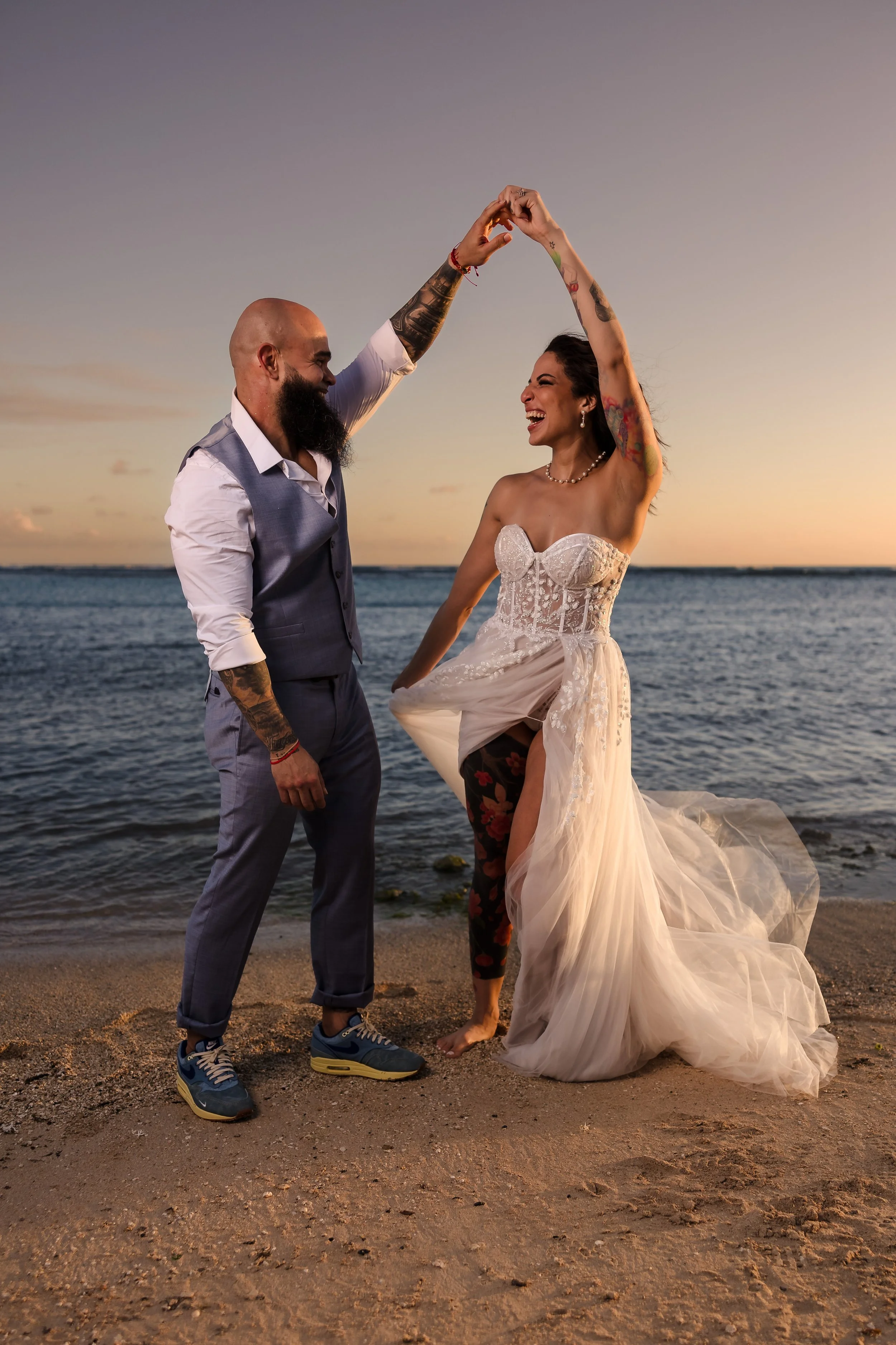 A newlywed couple dancing on a beach at sunset. The man has a bald head, beard, tattoos, and is wearing a vest and pants. The woman is wearing a strapless wedding dress, has tattoos and dark hair. They are smiling and holding hands aloft while standi