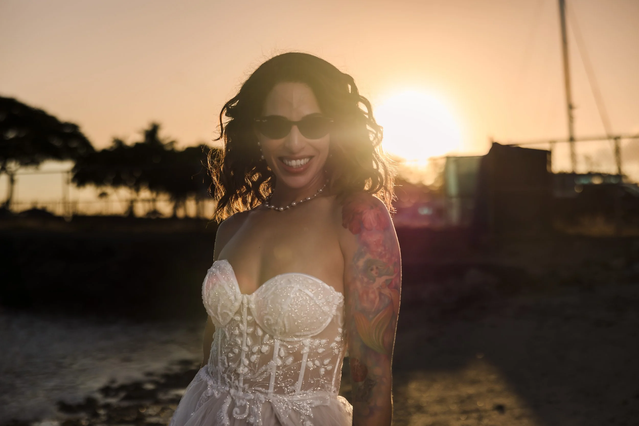 A woman in a wedding dress with tattoos on her arm, wearing sunglasses and a pearl necklace, standing on a beach at sunset.