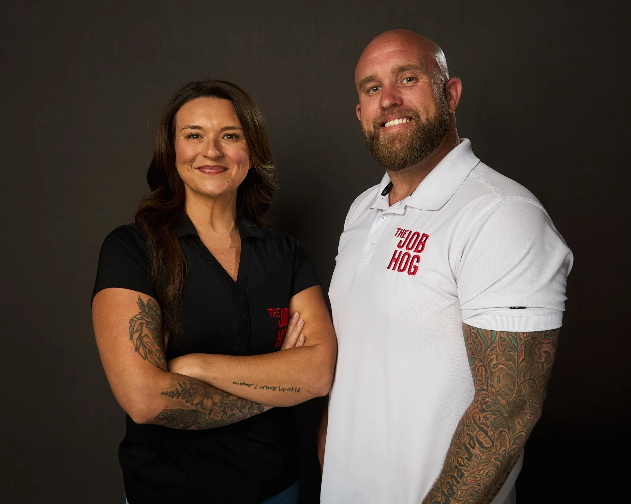 A woman with long brown hair and tattoos on her arms stands with her arms crossed, smiling. Next to her is a large, tattooed man with a beard, bald head, and a white polo shirt that reads 'The Job Hog'. They are against a dark background.