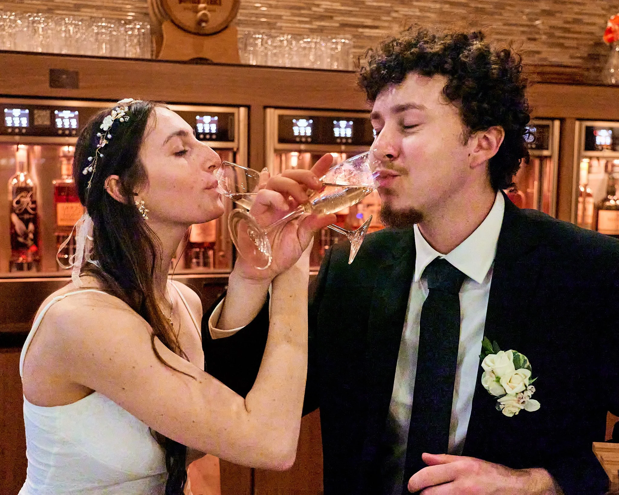 A bride and groom are toasting with champagne glasses in a warmly lit, wood-paneled room. The bride, with long dark hair wearing a floral headpiece and a white dress, has her eyes closed as she drinks. The groom, with curly dark hair and a beard, is 
