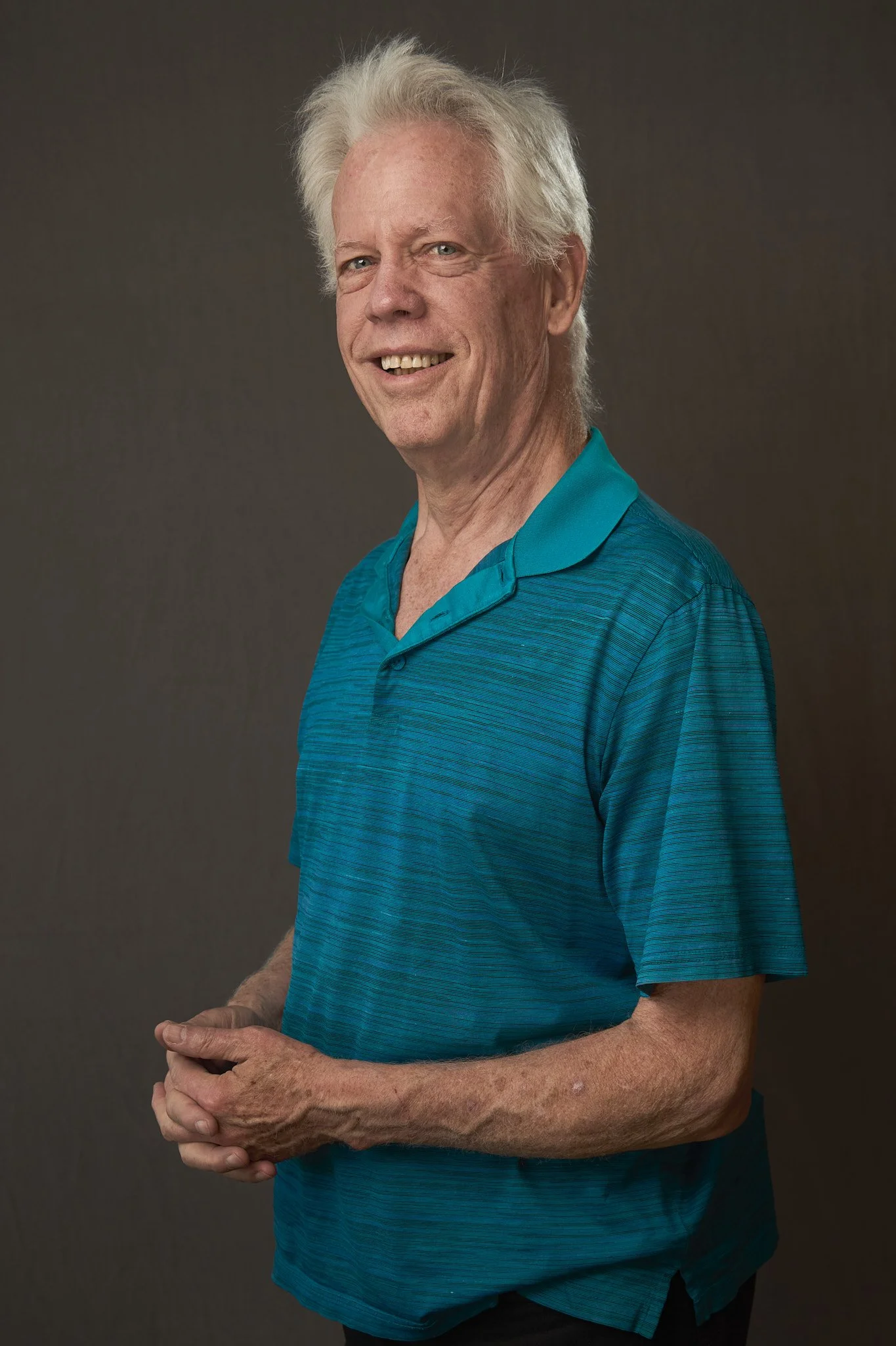 Portrait of an elderly man with white hair, smiling slightly, wearing a teal polo shirt, standing against a dark background.