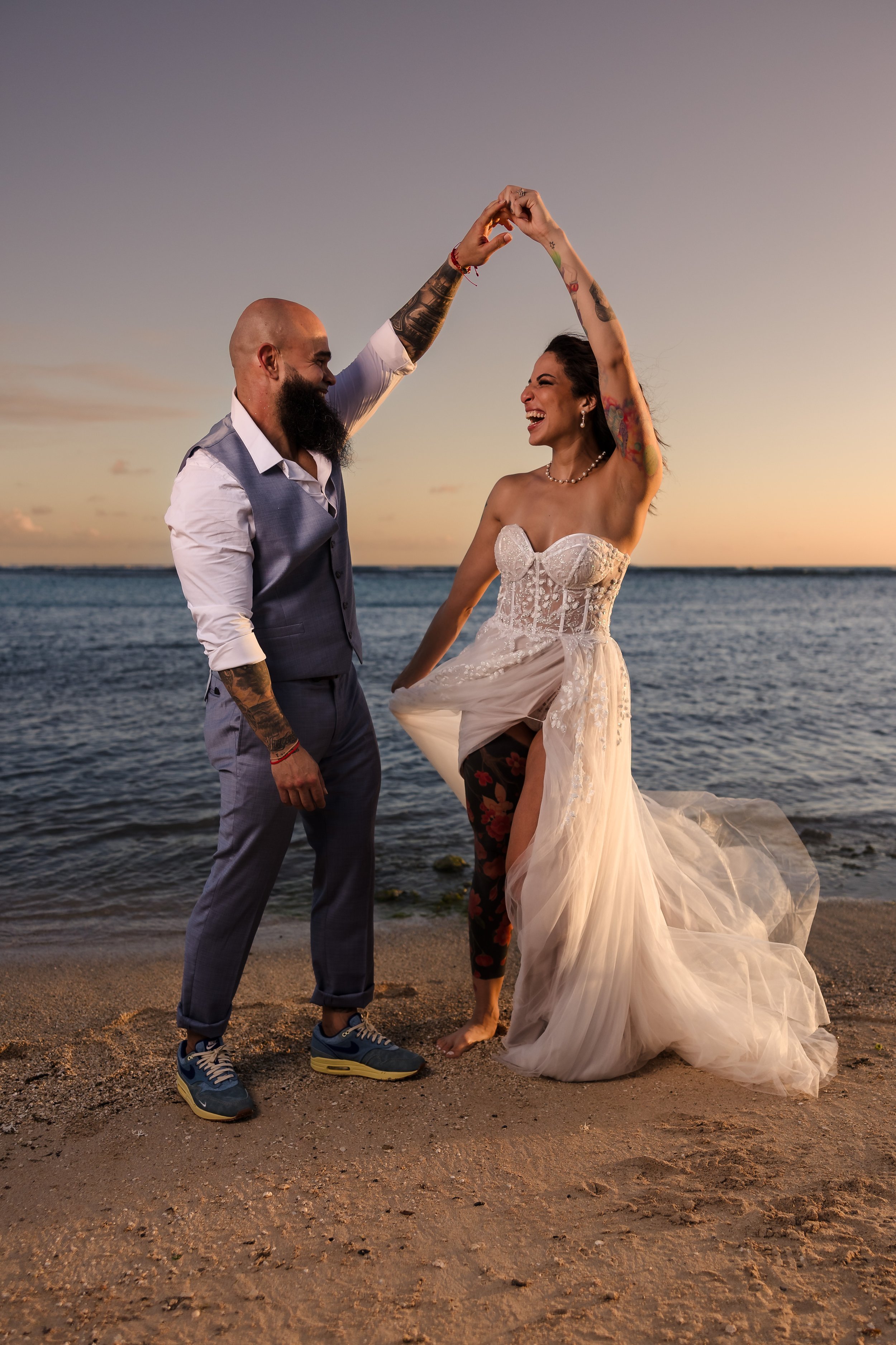 A happy couple on a beach at sunset, dancing and smiling, with the man wearing casual clothes and the woman in a wedding dress.