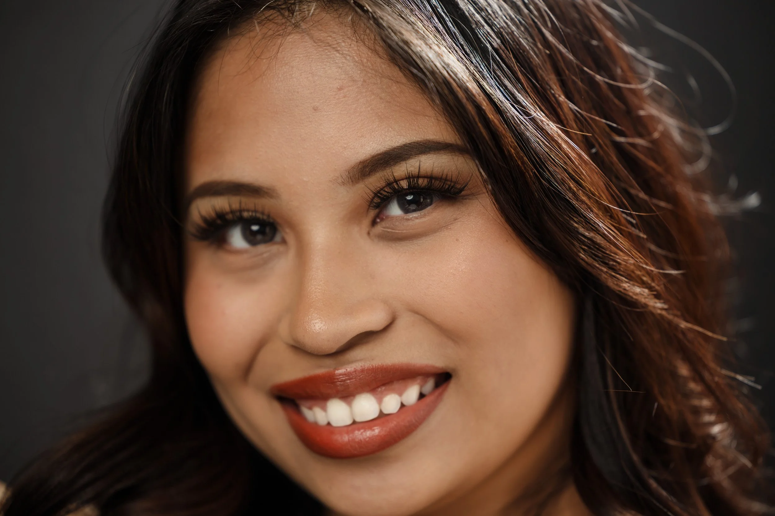 Close-up of a woman's face showing her smiling mouth, white teeth, and expressive eyes with long eyelashes. She has long, wavy hair and makeup with bold lipstick.