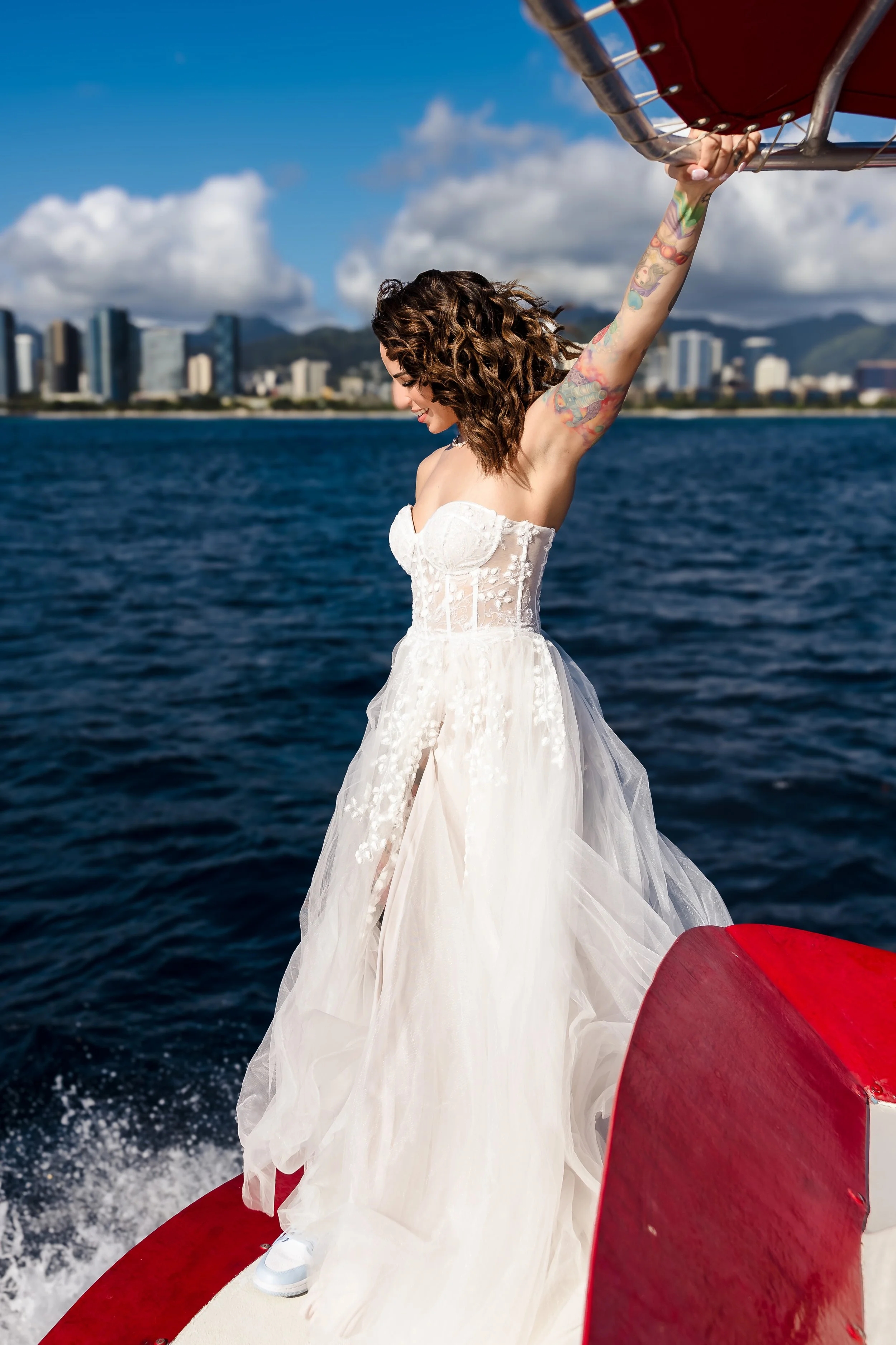 A woman wearing a white dress with tattoos on her arm is holding onto a boat's railing while on the water, with a city skyline and mountains in the background.