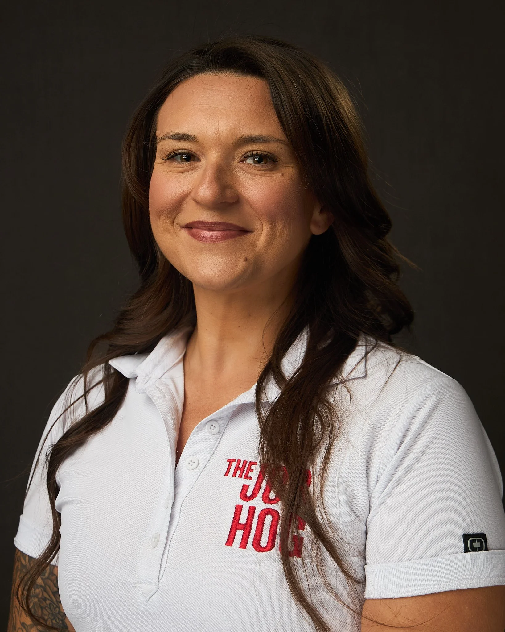A woman smiling, wearing a white polo shirt with red text on it, and has long wavy brown hair.