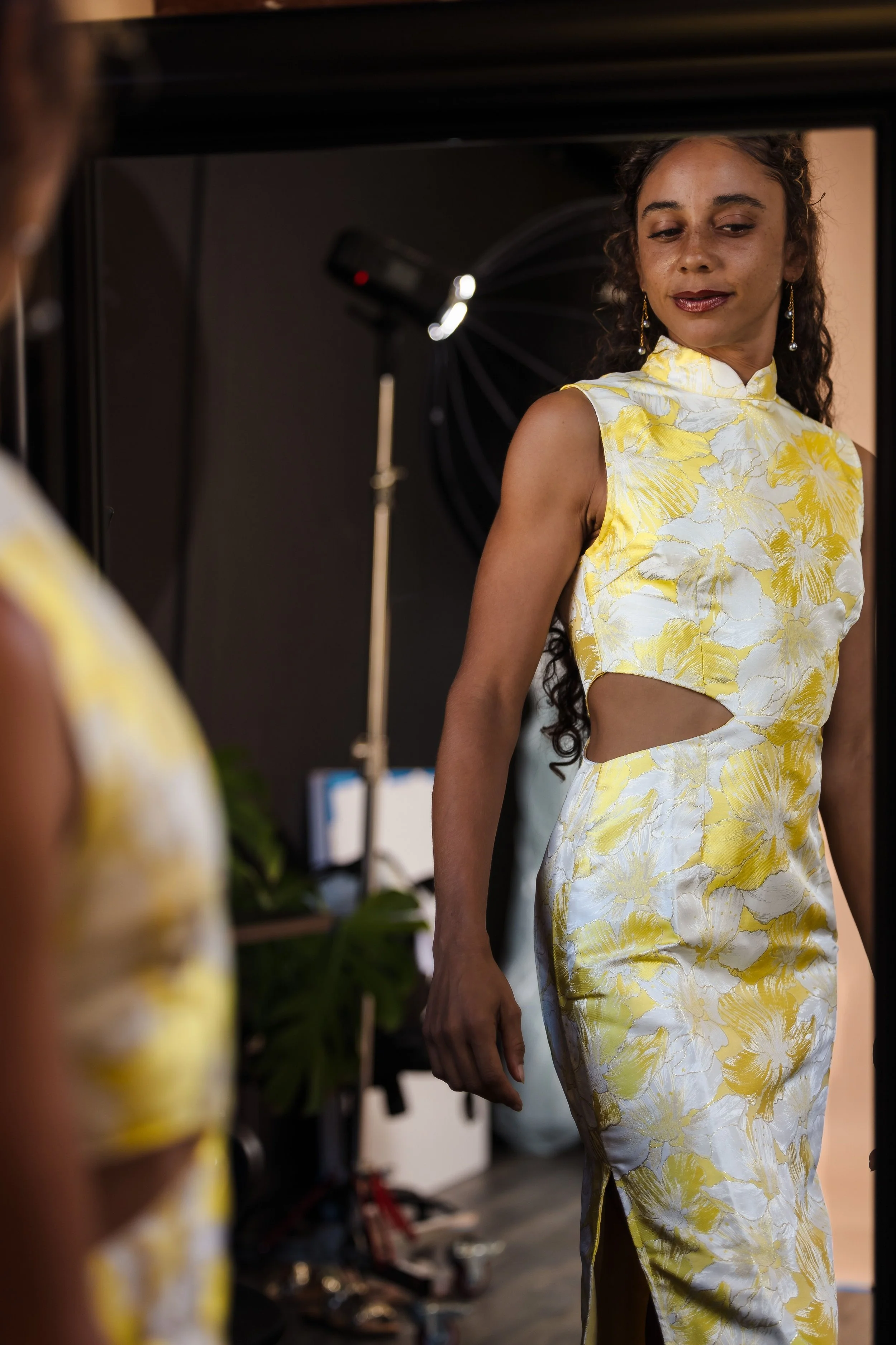 A woman with curly hair wearing a yellow floral dress looking at her reflection in a mirror.