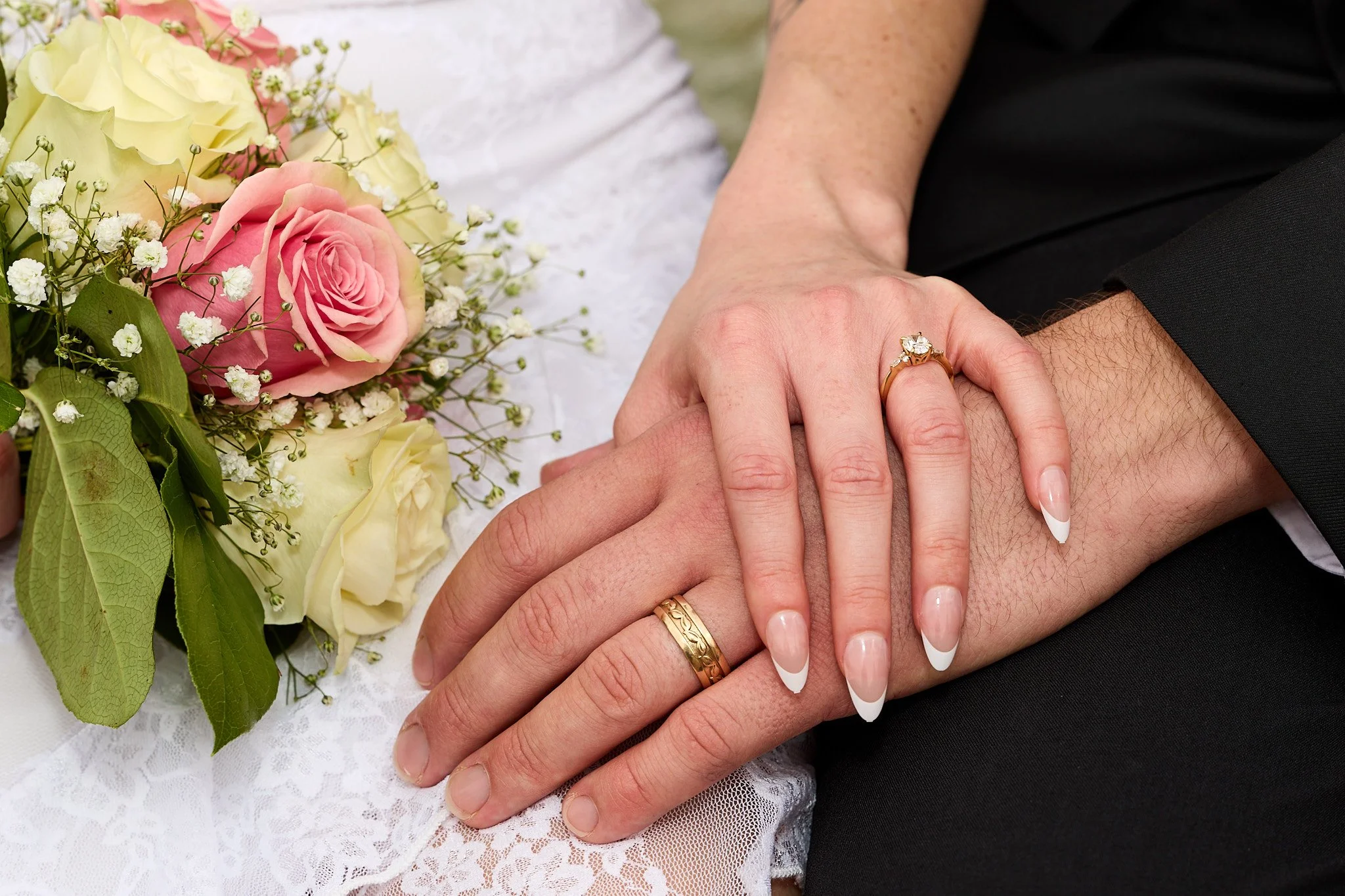 Close-up of newlyweds' hands resting on white lace fabric, with wedding rings, near a bouquet of pink and cream roses with baby's breath and green leaves.