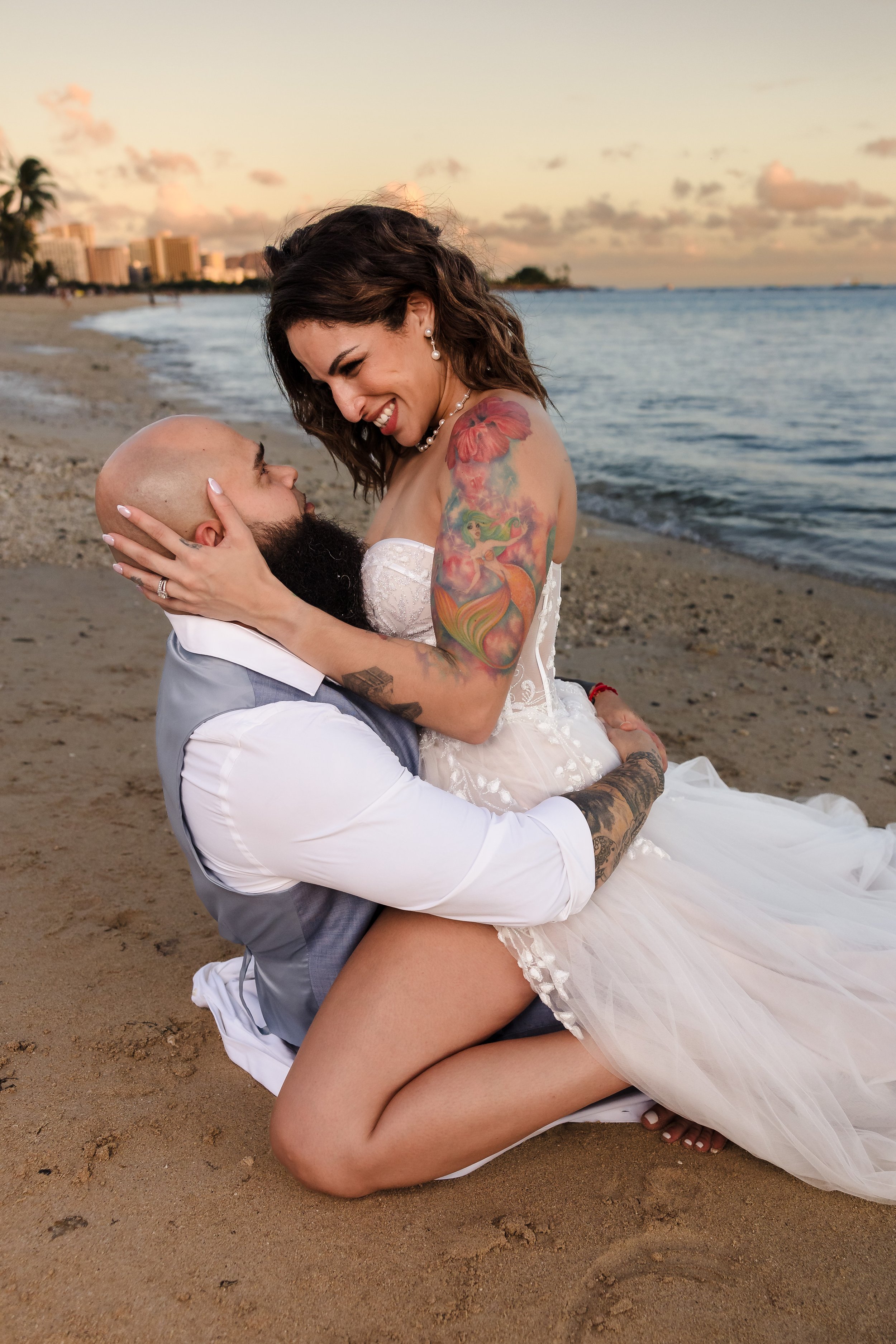 A couple on the beach, the woman wearing a wedding dress and the man in a vest and shirt, holding each other and smiling as they look at each other with the ocean and city skyline in the background during sunset.