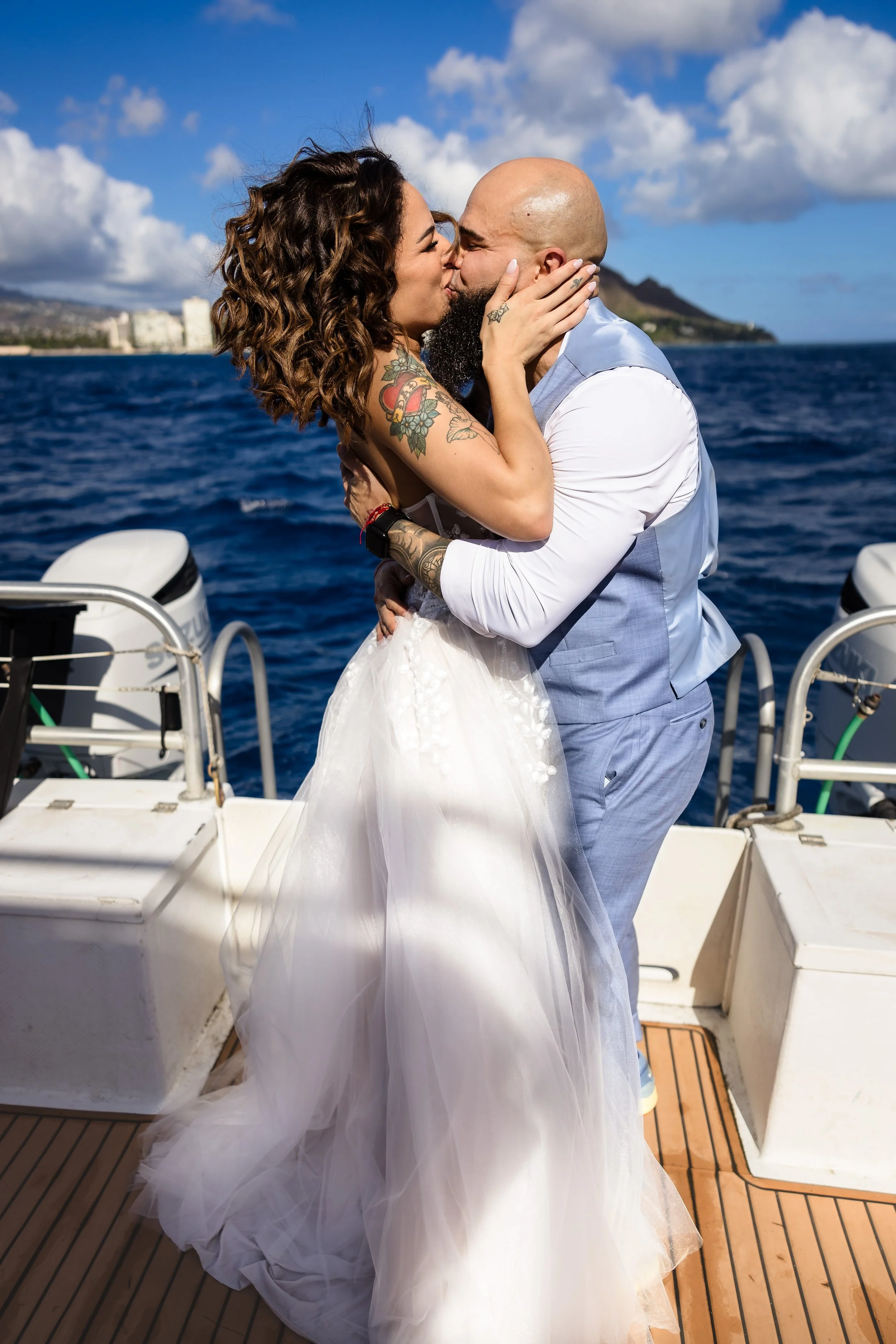A couple sharing a kiss on a boat, with the ocean, a distant shoreline, and a partly cloudy sky in the background.