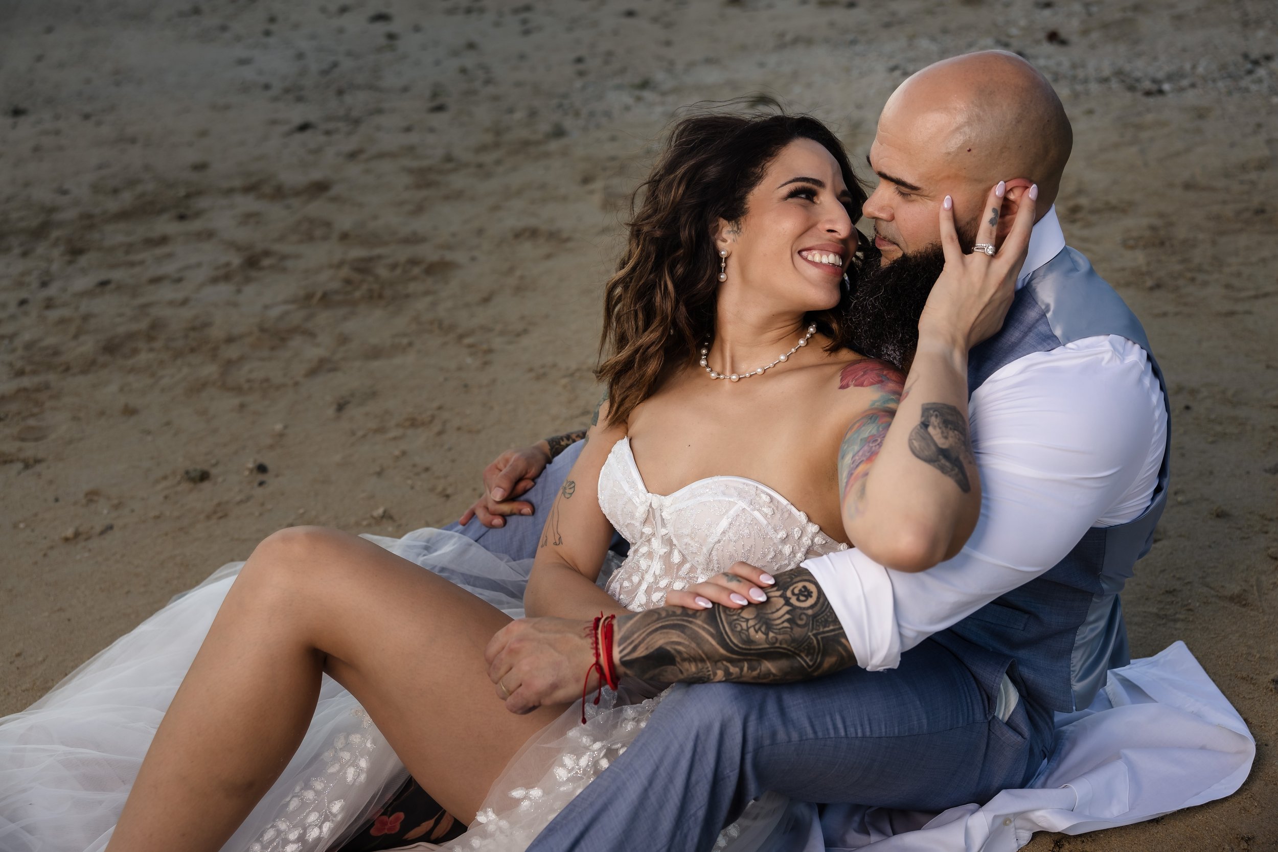 A couple on the beach, smiling and embracing. The woman is wearing a white dress and jewelry, and the man is in a white shirt and gray vest. They are sitting on the sand, close together, with the woman touching the man's face.