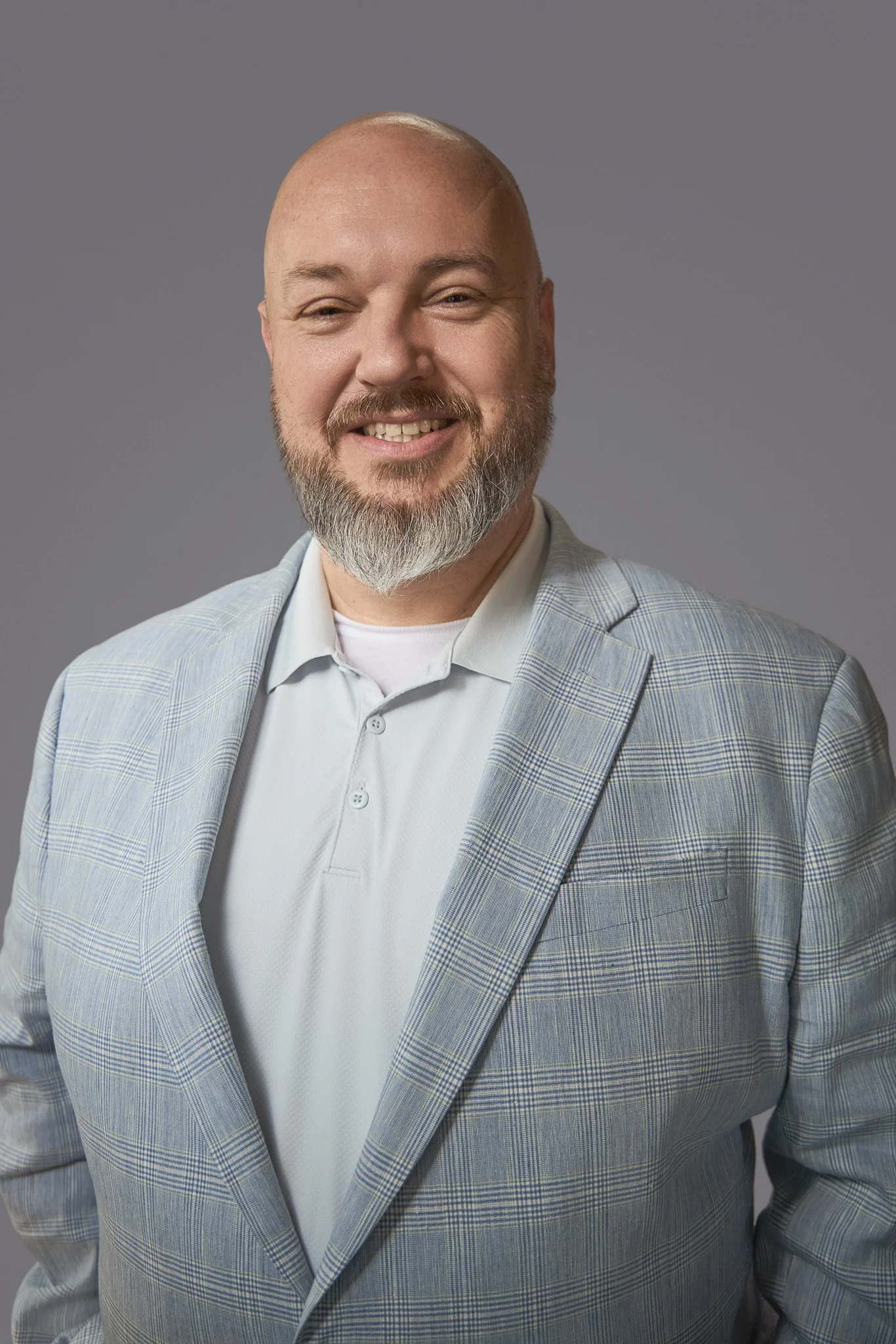A bald man with a beard smiling while wearing a light plaid suit jacket and a light gray polo shirt against a gray background.