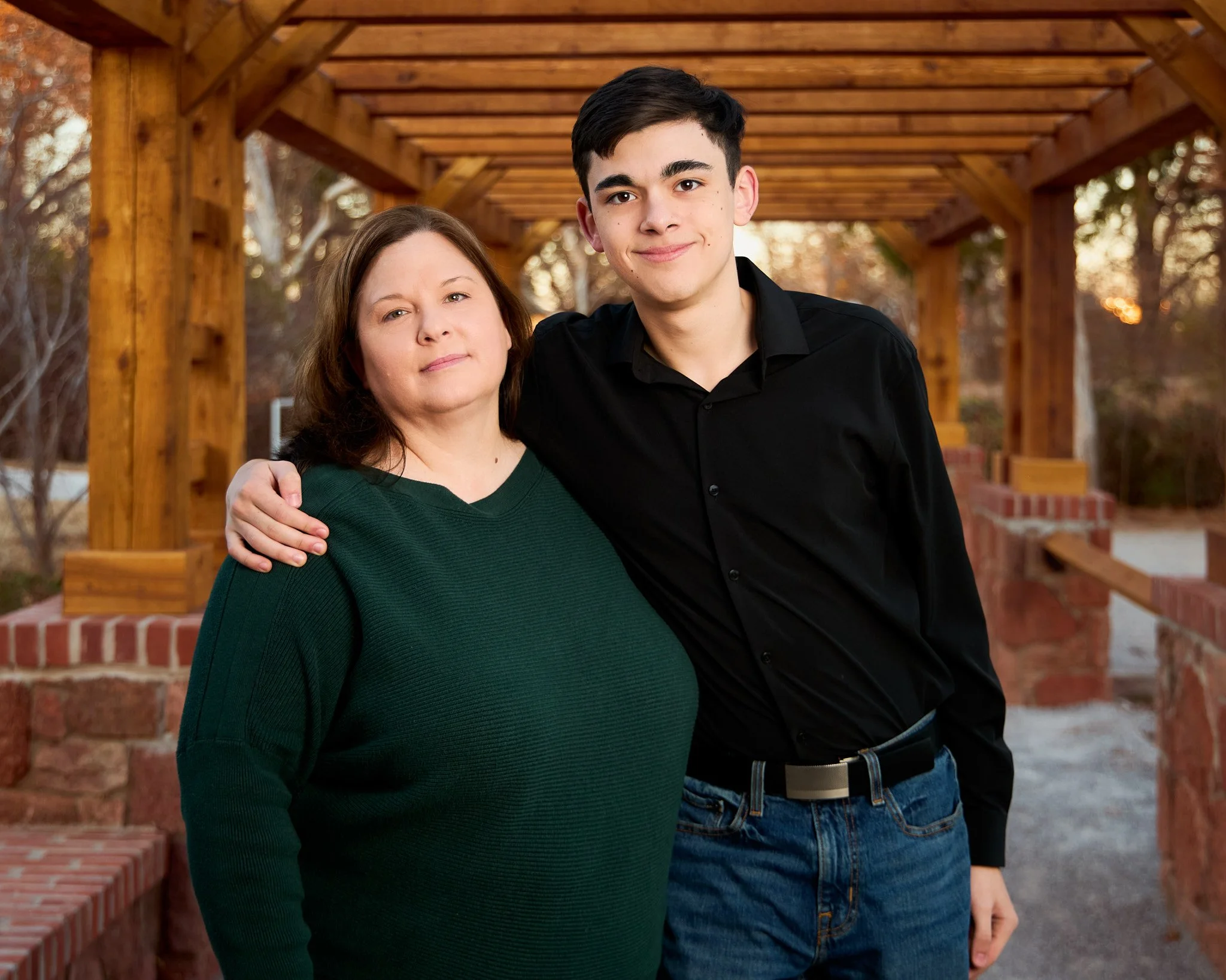 A woman and a teenage boy standing close together outdoors under a wooden pergola, with the woman having her arm around the boy's shoulder, both smiling softly at the camera, autumn trees in the background.