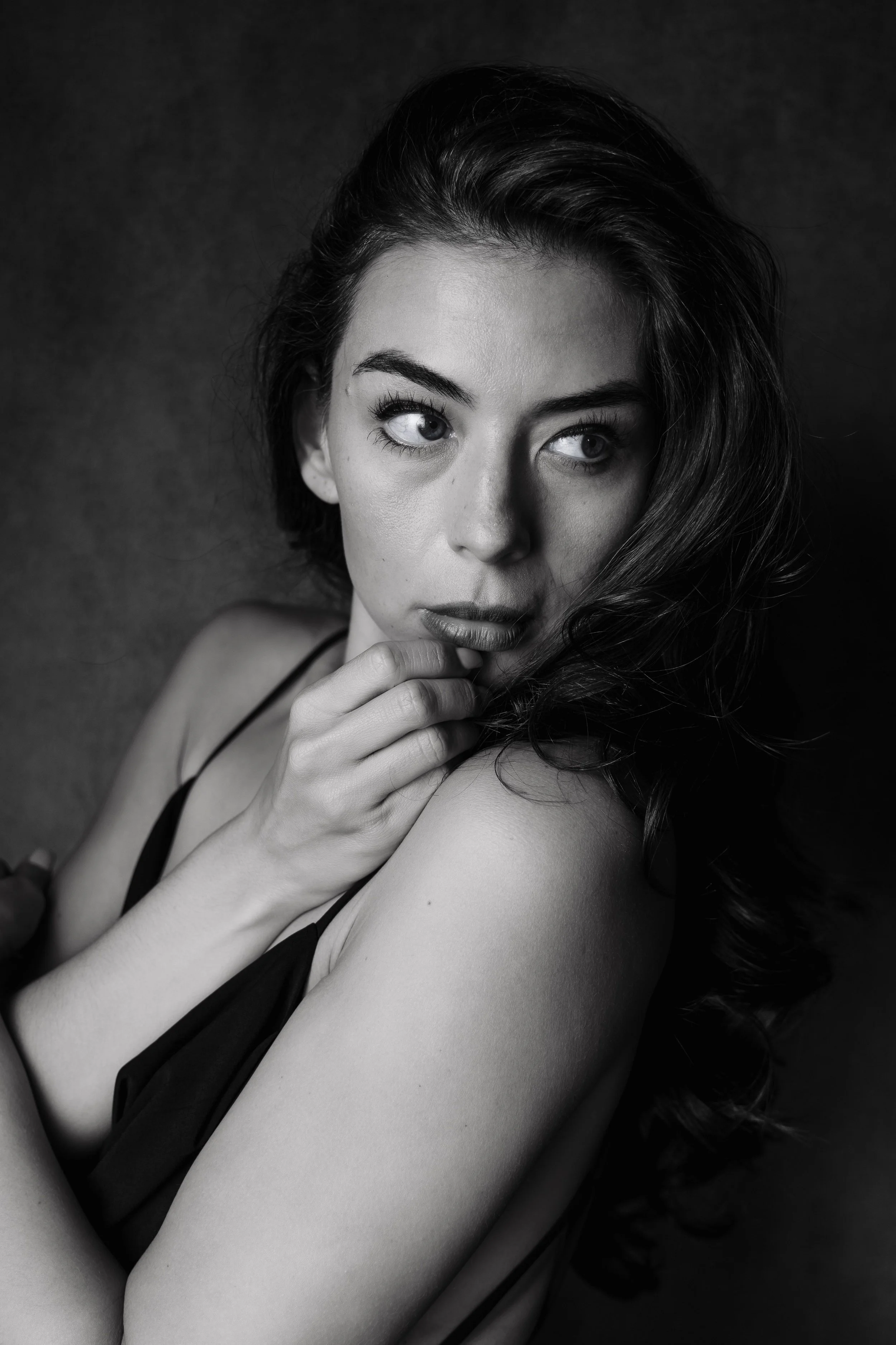 Black and white portrait of a young woman with wavy hair, looking slightly upwards with her hand near her chin, wearing a spaghetti strap top.