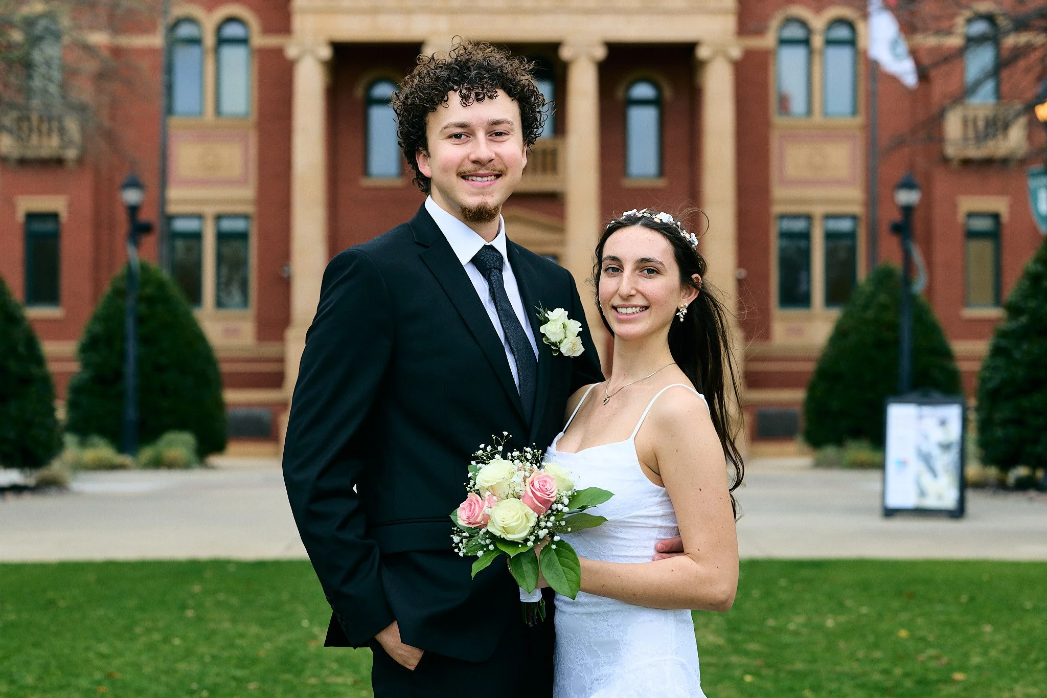 A smiling bride and groom standing outdoors in front of a historic red brick building, wearing wedding attire with the bride holding a bouquet of white and pink roses.