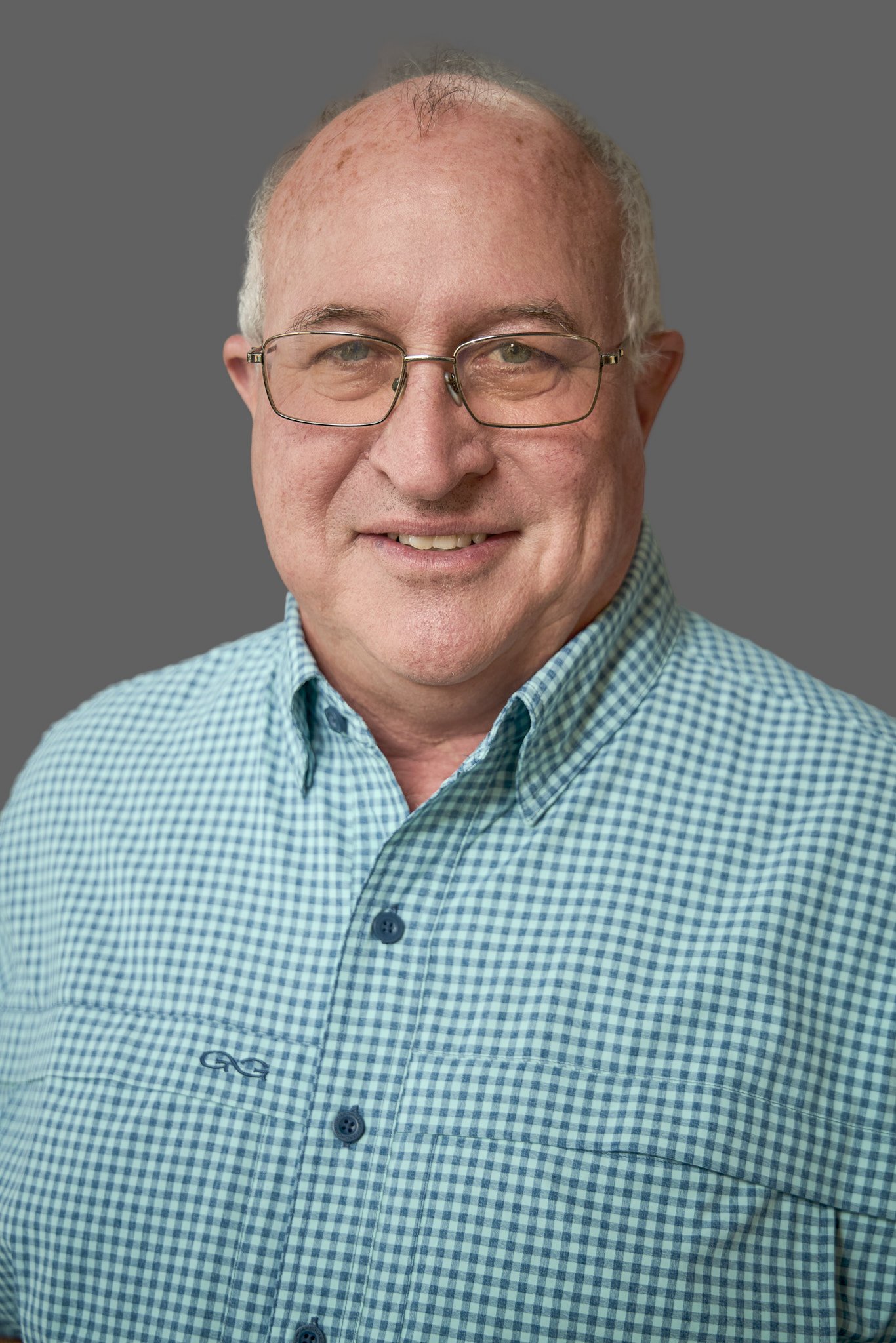 Portrait of an older man wearing glasses and a blue checkered shirt, smiling, against a gray background.