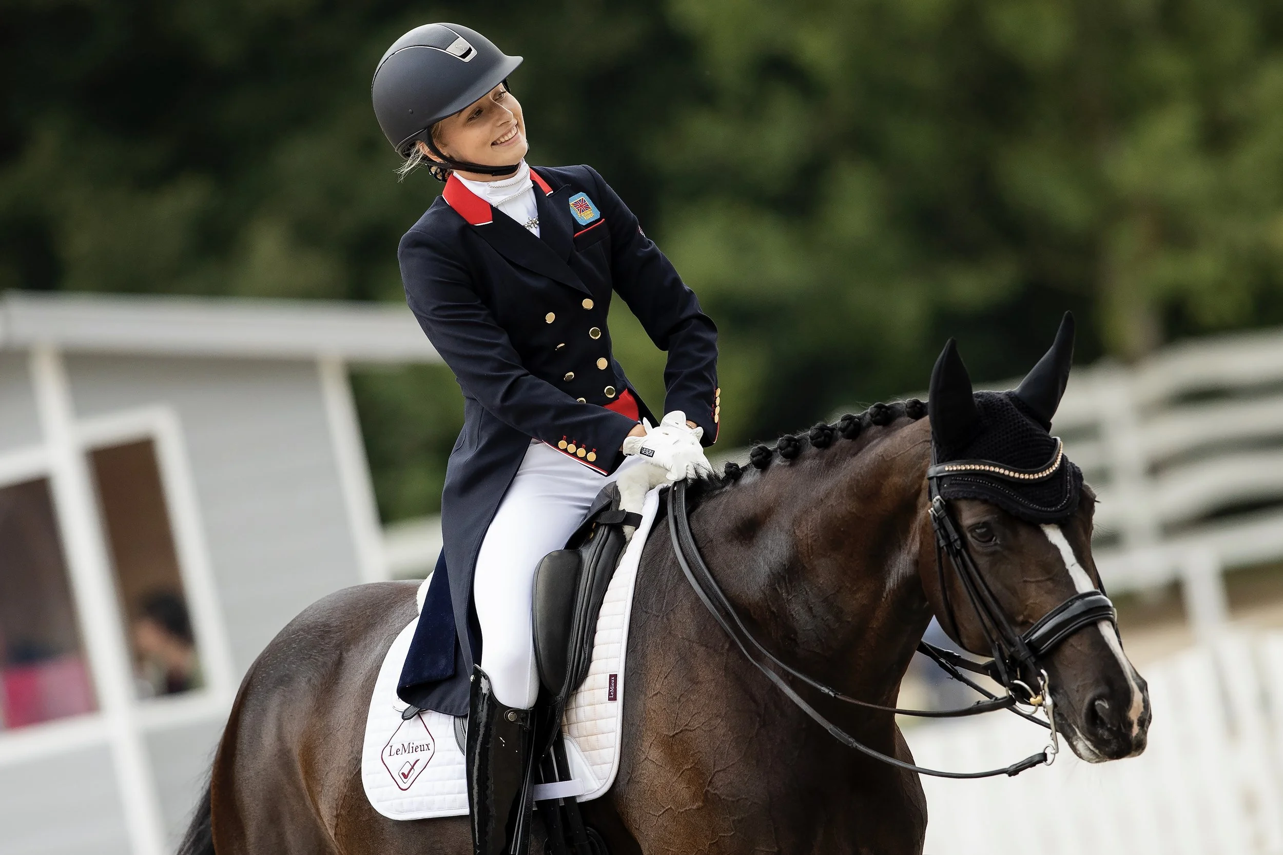 A woman dressed in equestrian riding attire, including a navy jacket, white riding pants, and a helmet, sitting on a brown horse with a black mane and a decorative ear bonnet, smiling during a riding event.
