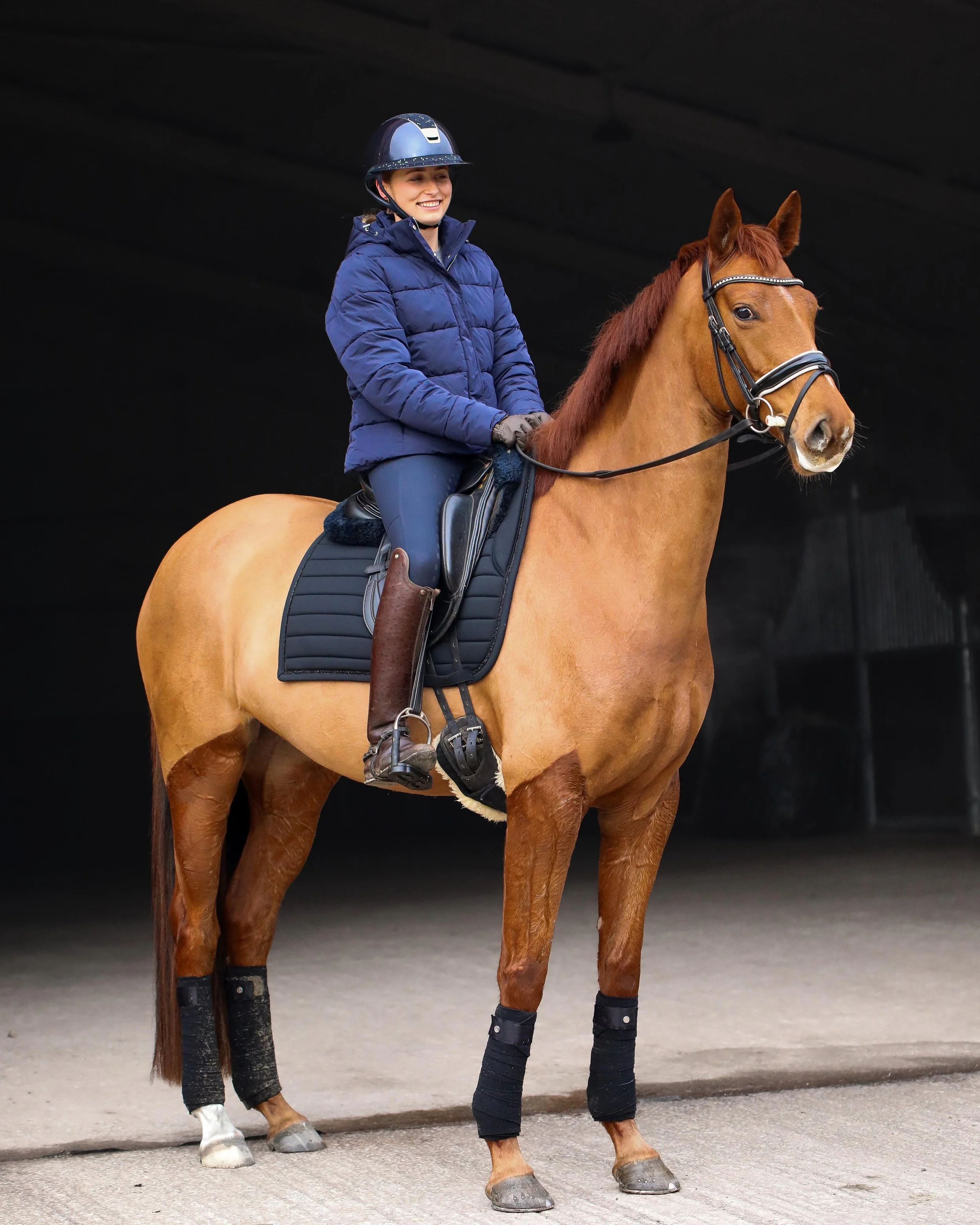 A woman riding a chestnut horse in an indoor riding arena, wearing a blue jacket, riding helmet, and riding boots.
