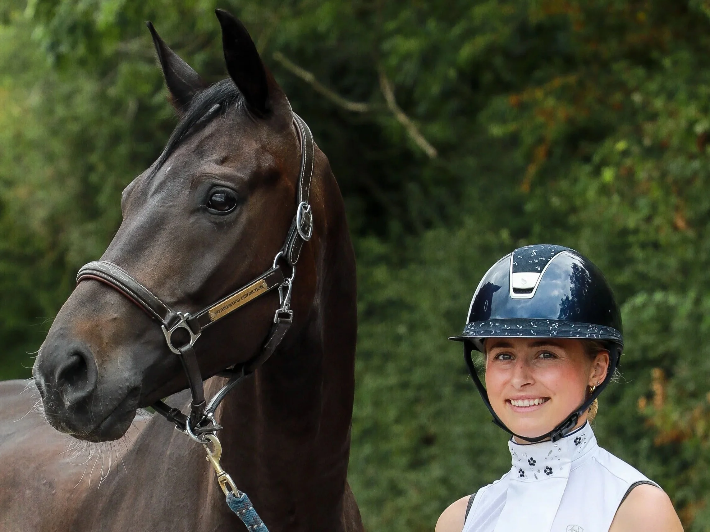 A smiling woman in a riding helmet standing next to a dark brown horse outdoors with green trees in the background.