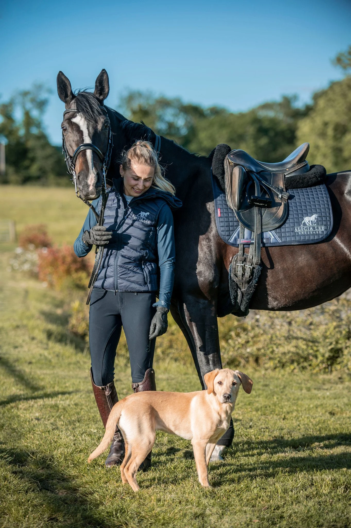 A woman standing next to a black horse with saddle and bridle, holding the horse's reins, while a tan dog stands on the grass closer to the camera in an outdoor setting with trees and bushes in the background.