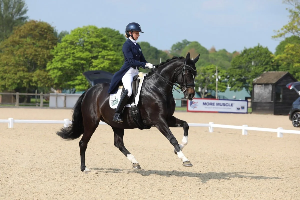 A female equestrian wearing formal riding attire, including a helmet, jacket, and breeches, riding a dark brown horse with white markings on its legs in a dressage arena.