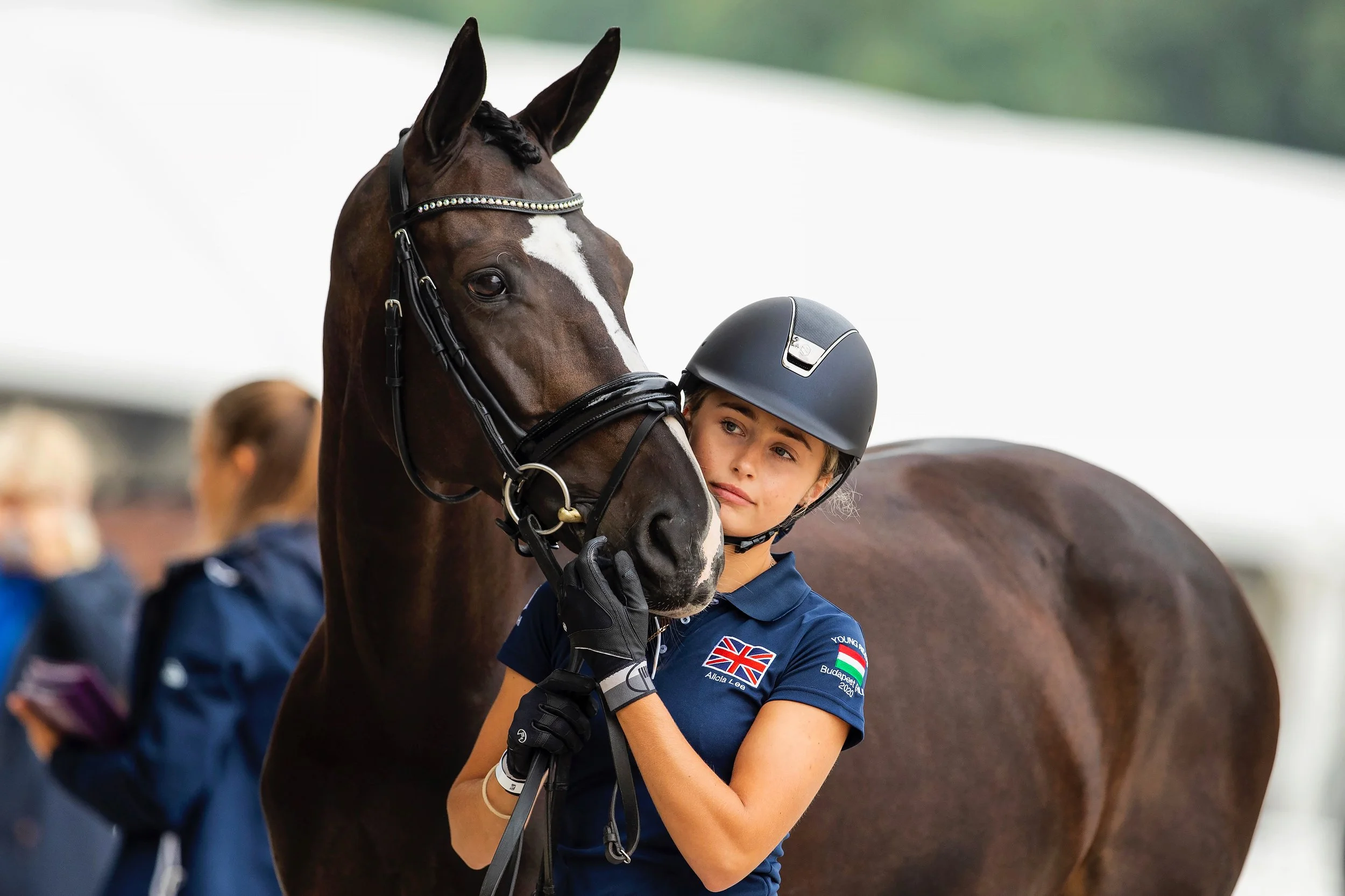 A young female equestrian athlete in a navy uniform with British flag and Hungarian flag patches on sleeves, wearing a black riding helmet, stands next to a dark brown horse with a white star on its forehead and a bridle. She holds the horse's reins 