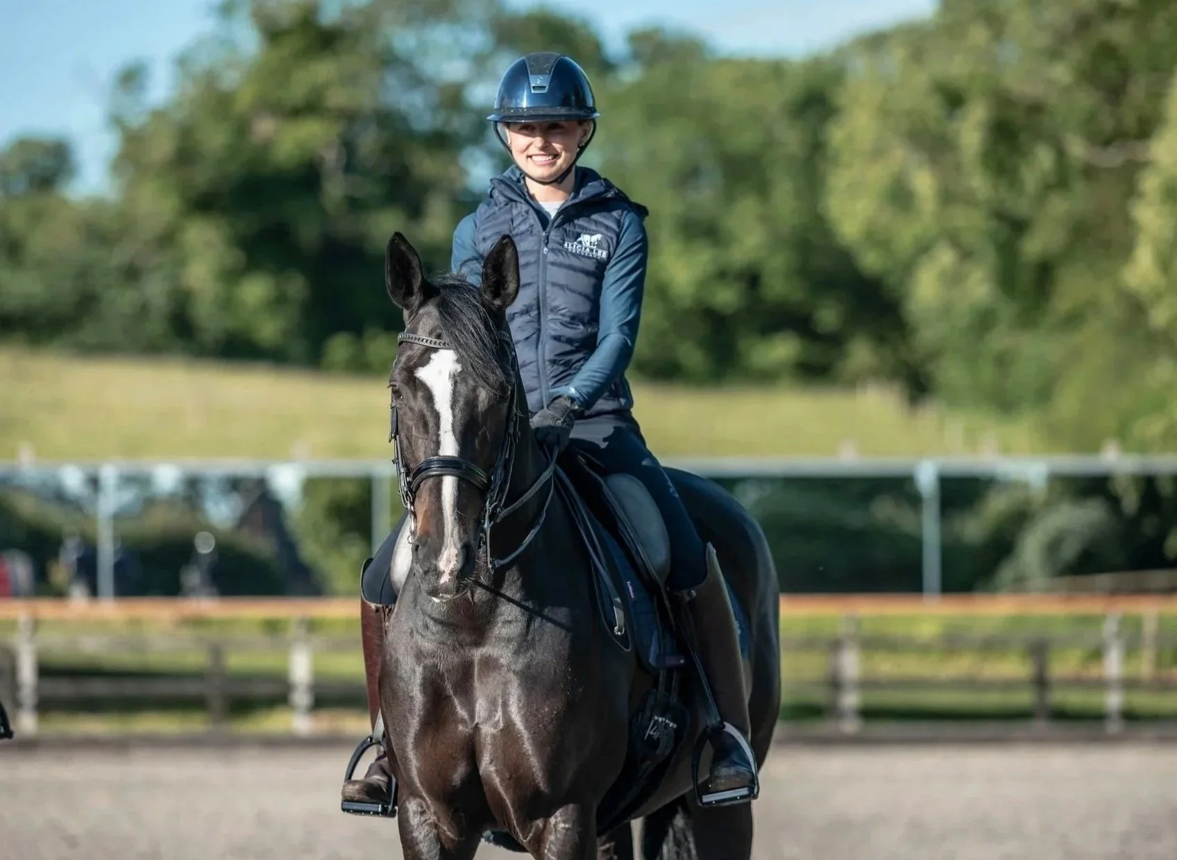 A young woman riding a dark gray horse outdoors on a sunny day, wearing a helmet, blue jacket, and gloves, with a green blurred background of trees.