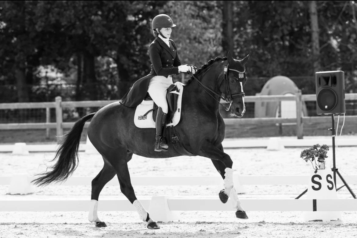 A woman riding a horse in an equestrian event, wearing a helmet and riding attire, with a speaker and flower arrangement in the background.