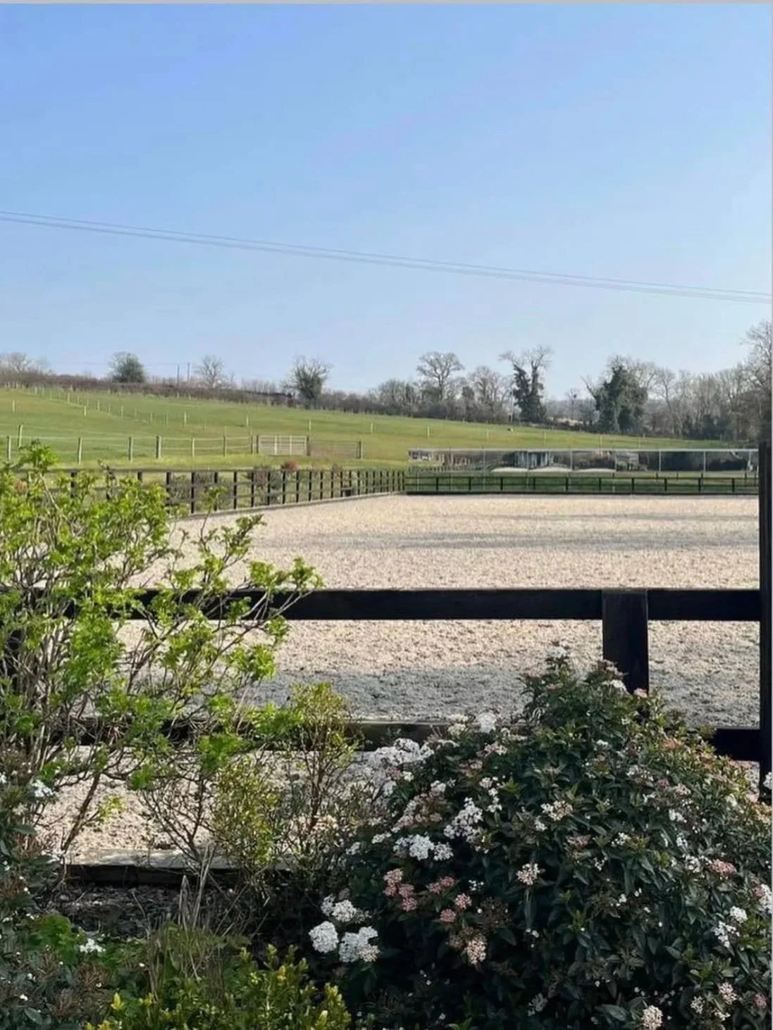 View of a horse riding arena with a sandy surface, surrounded by green hills and trees, and a clear blue sky.