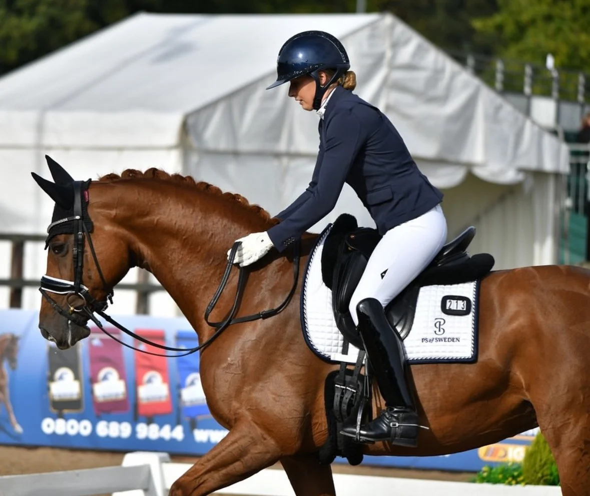 A female equestrian rider in a navy jacket, white riding pants, and a helmet, riding a chestnut horse during a competition, with tents and banners in the background.