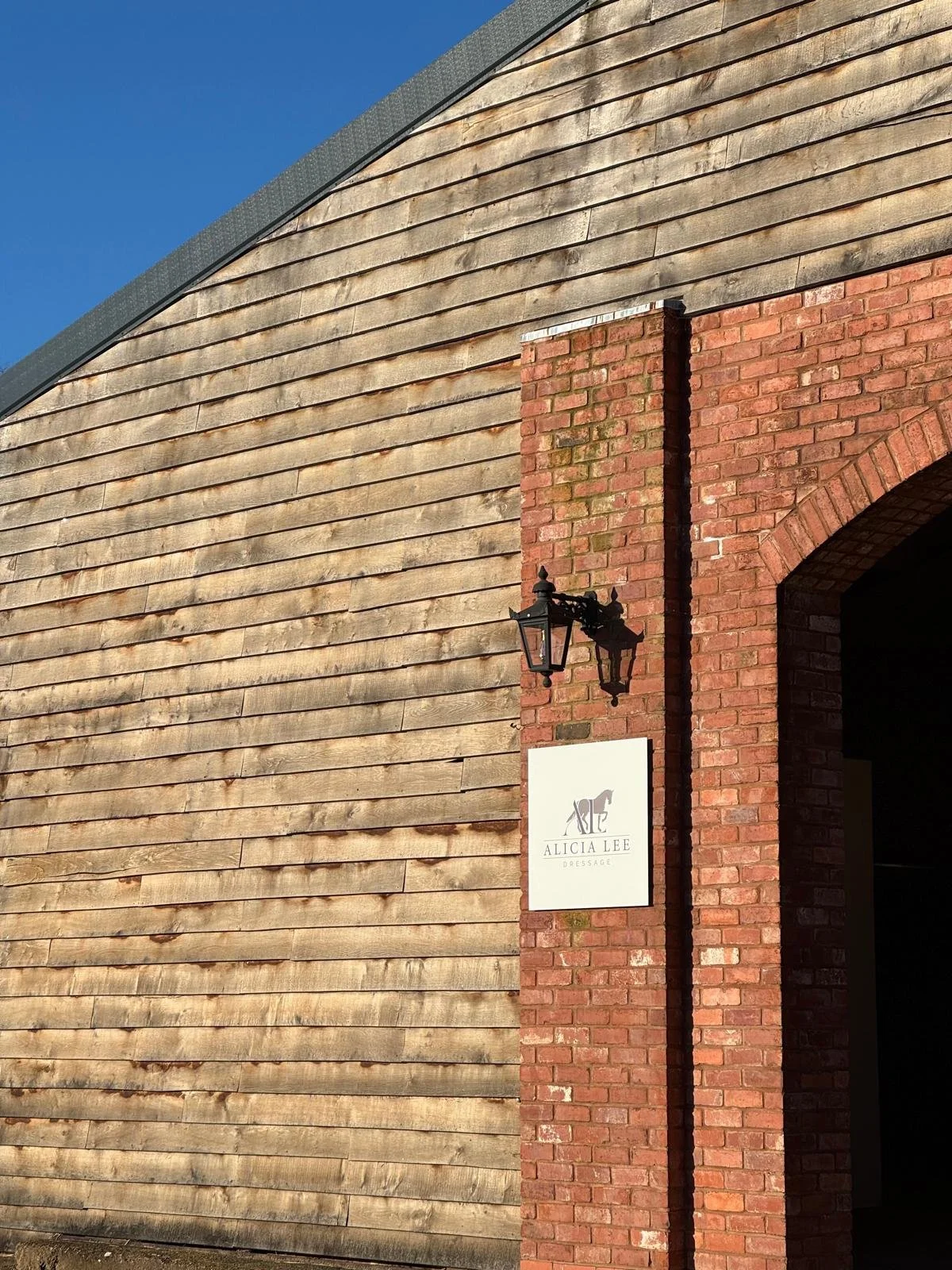 Exterior of a building with wooden siding and a brick archway entrance, featuring a black lantern and white sign with a horse logo and text reading 'Alicia Lee Dressage'.