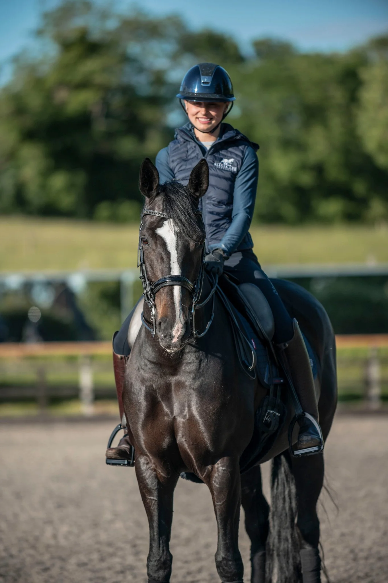 A woman riding a dark brown horse outdoors on a sunny day, wearing a helmet and riding gear, with trees in the background.