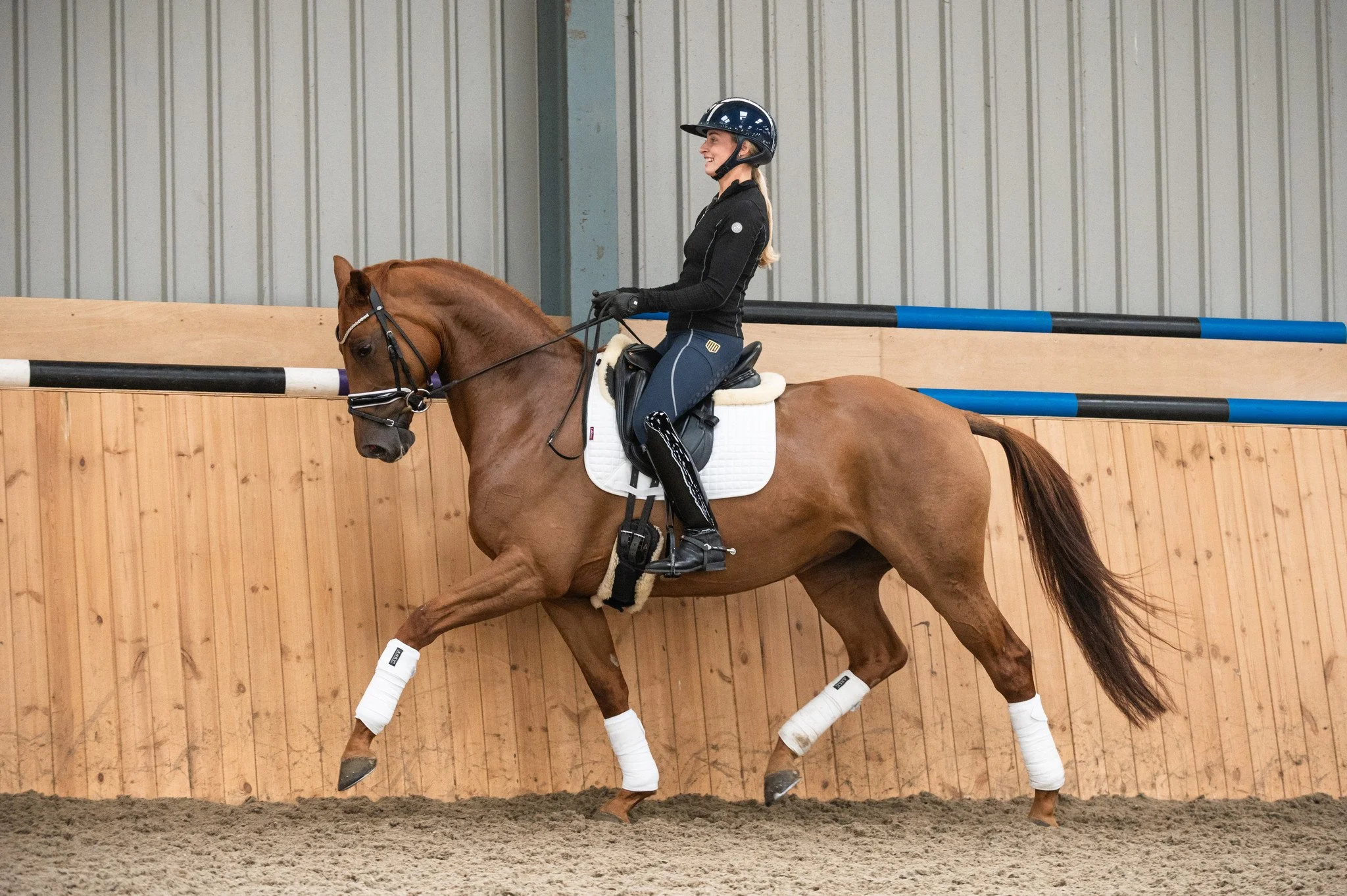 A woman in riding gear riding a chestnut horse inside an indoor riding arena.