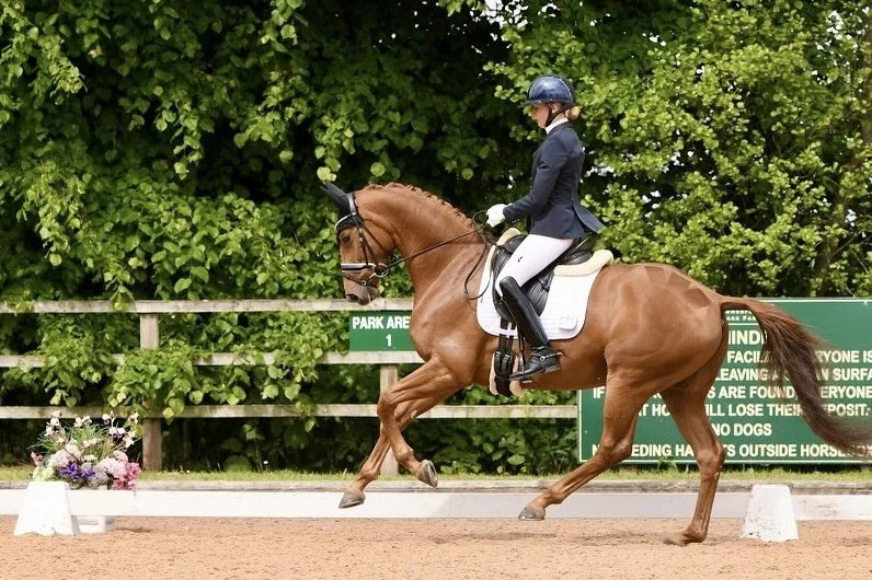 A woman riding a chestnut horse during a dressage competition at an outdoor arena with green foliage in the background.