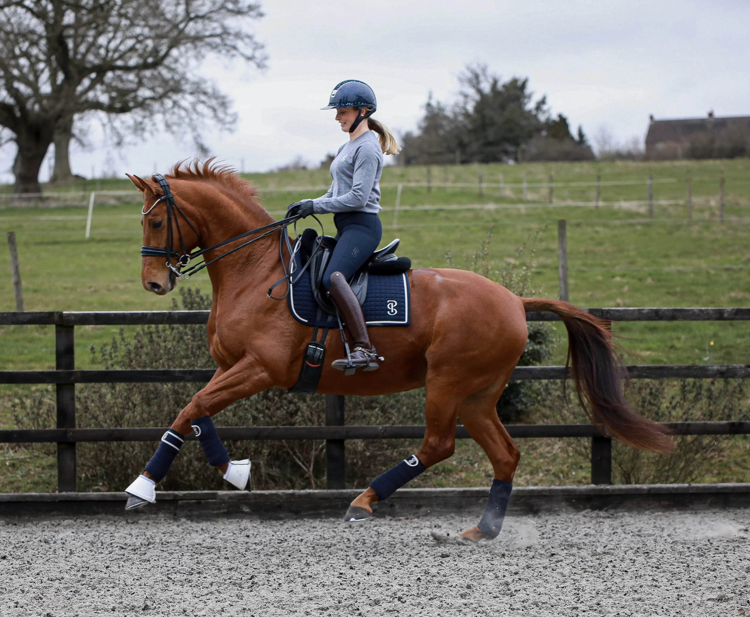 A woman riding a chestnut horse on an outdoor riding arena, wearing a helmet, grey sweater, dark riding pants, and riding boots, with black gloves and protective leg wraps on the horse.