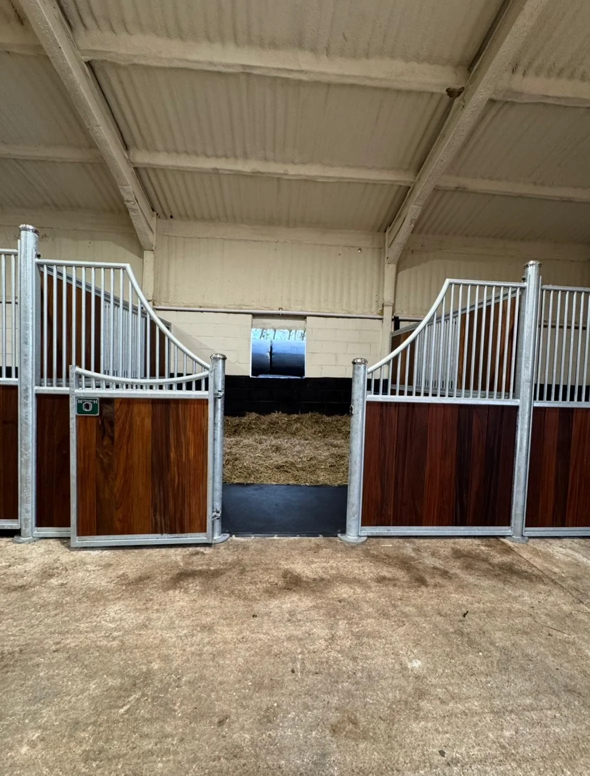 A horse stable with wooden and metal railings, hay on the floor, and a small window at the back, inside a barn with a white corrugated metal ceiling.
