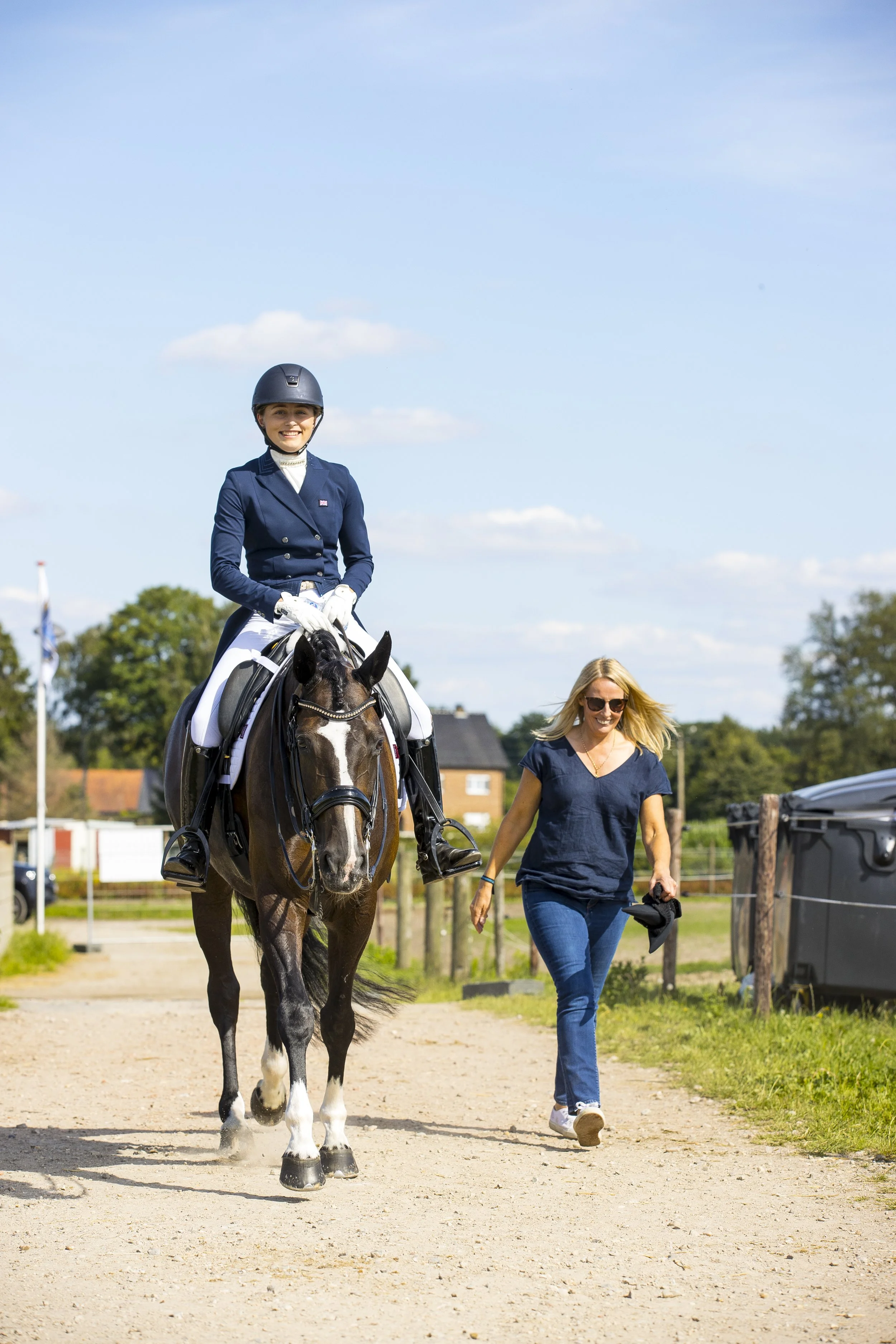 A woman walking beside a female horseback rider in formal riding attire on a dirt path outdoors under a blue sky