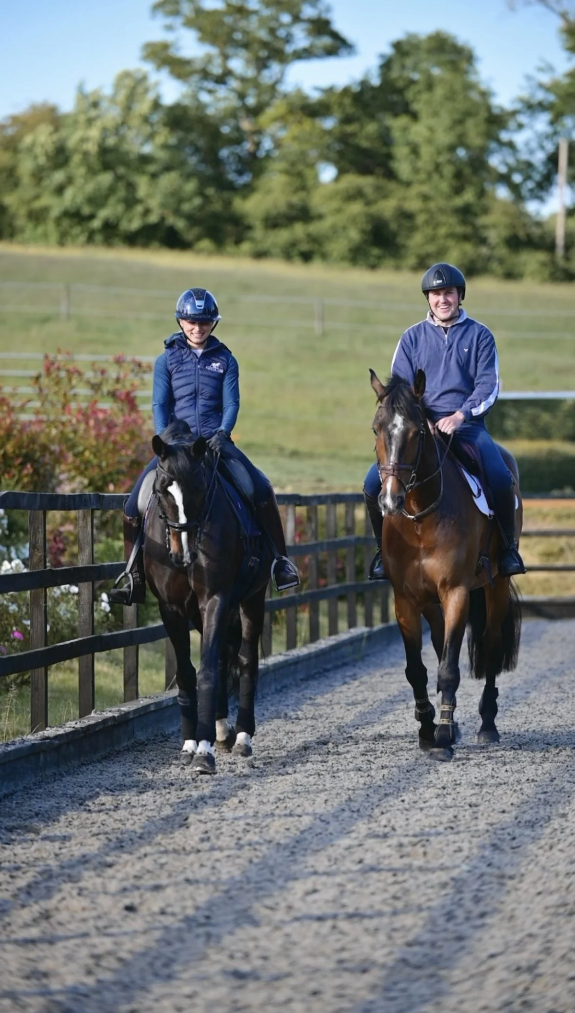 Two people riding horses on a dirt path in an outdoor riding arena with a fence, trees, and blue sky in the background.