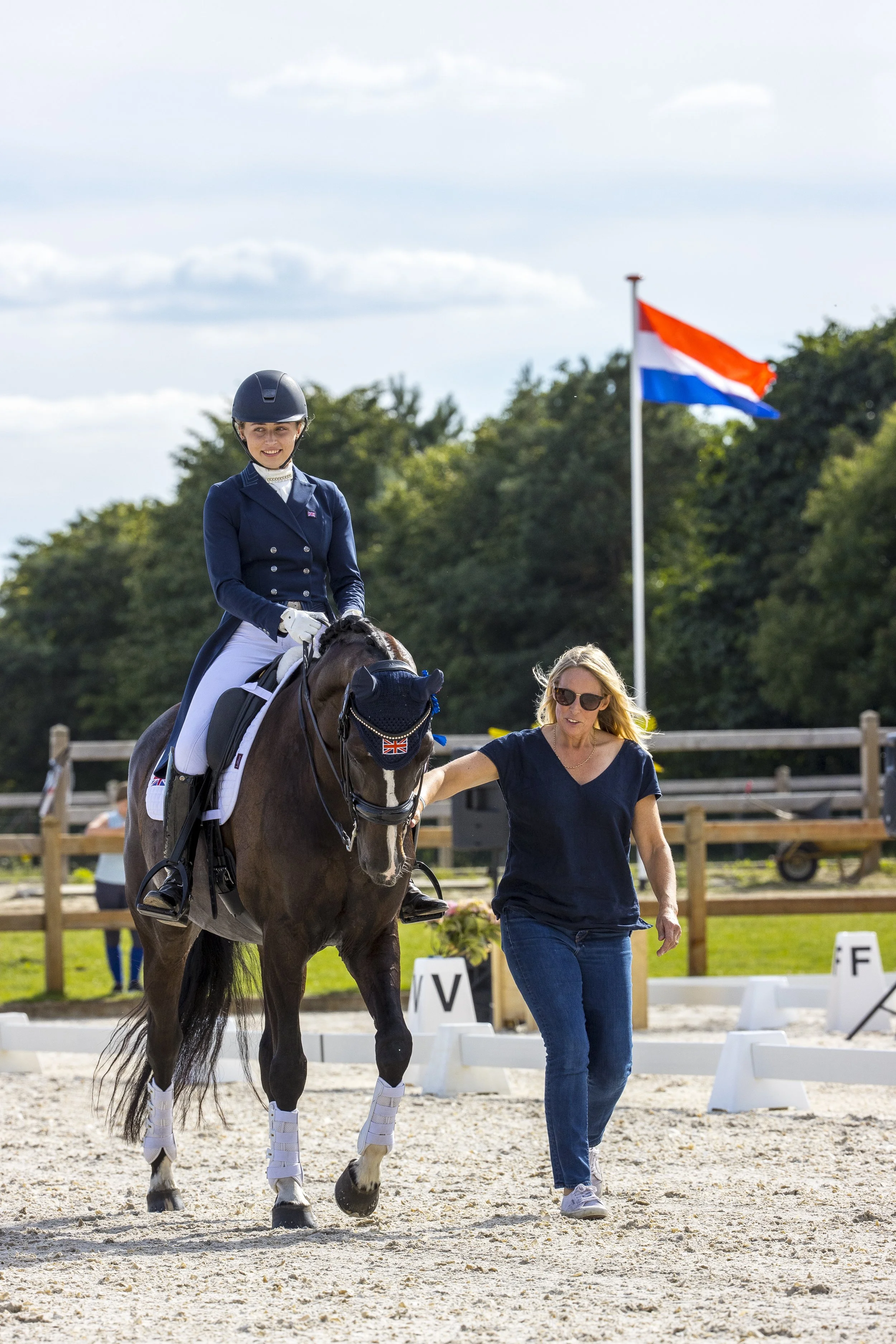 A woman riding a horse in an outdoor arena with a Dutch flag and trees in the background, her hair being blown by the wind.