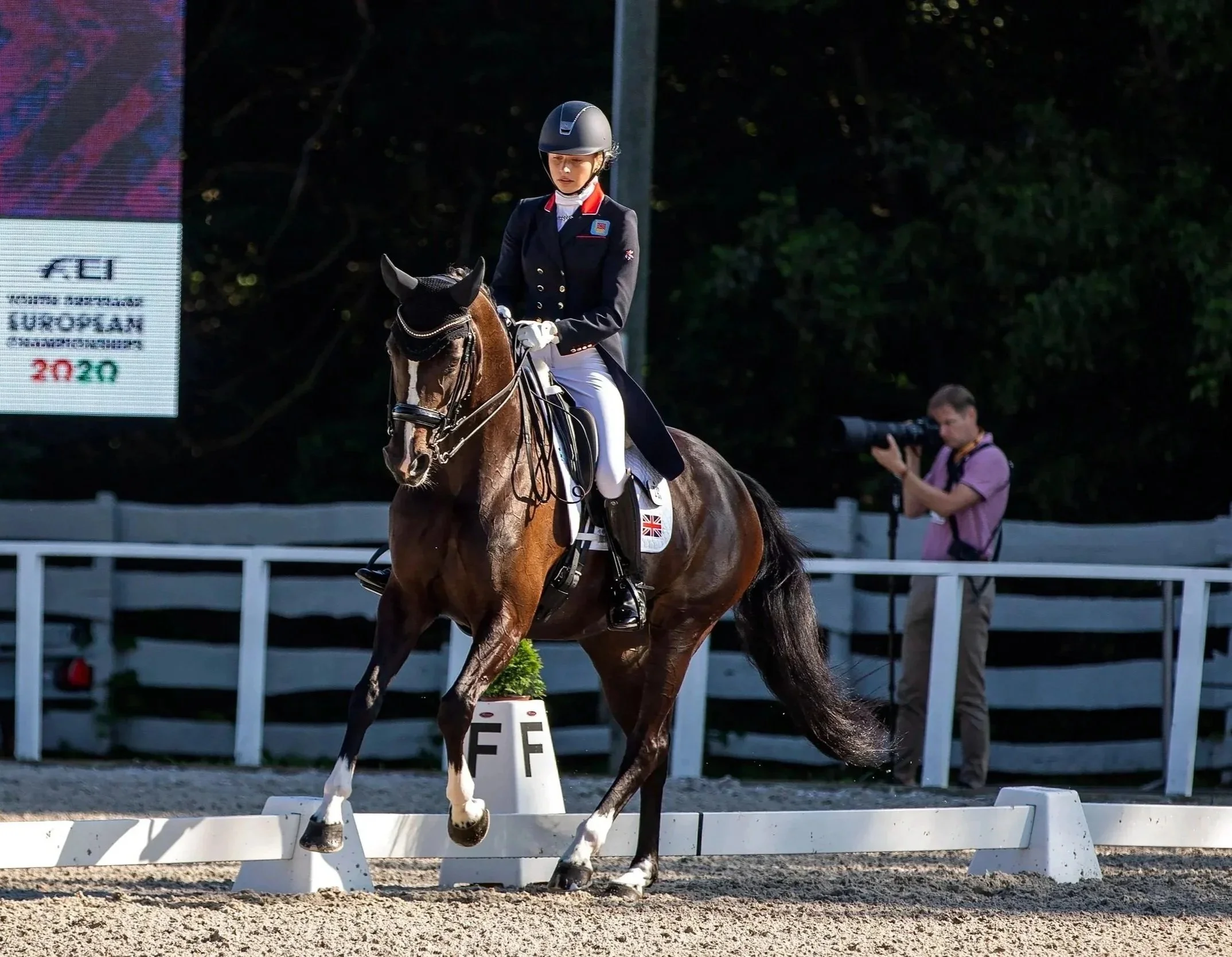 Equestrian rider in formal attire riding a brown horse in a dressage event with a photographer capturing the moment in the background.