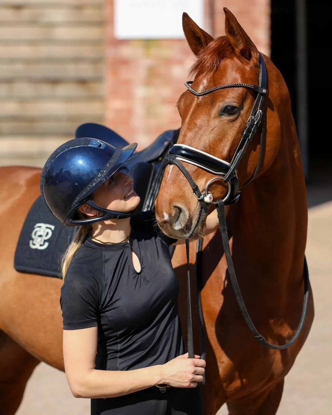 A woman wearing a helmet stands next to a brown horse and hugs its face, smiling.