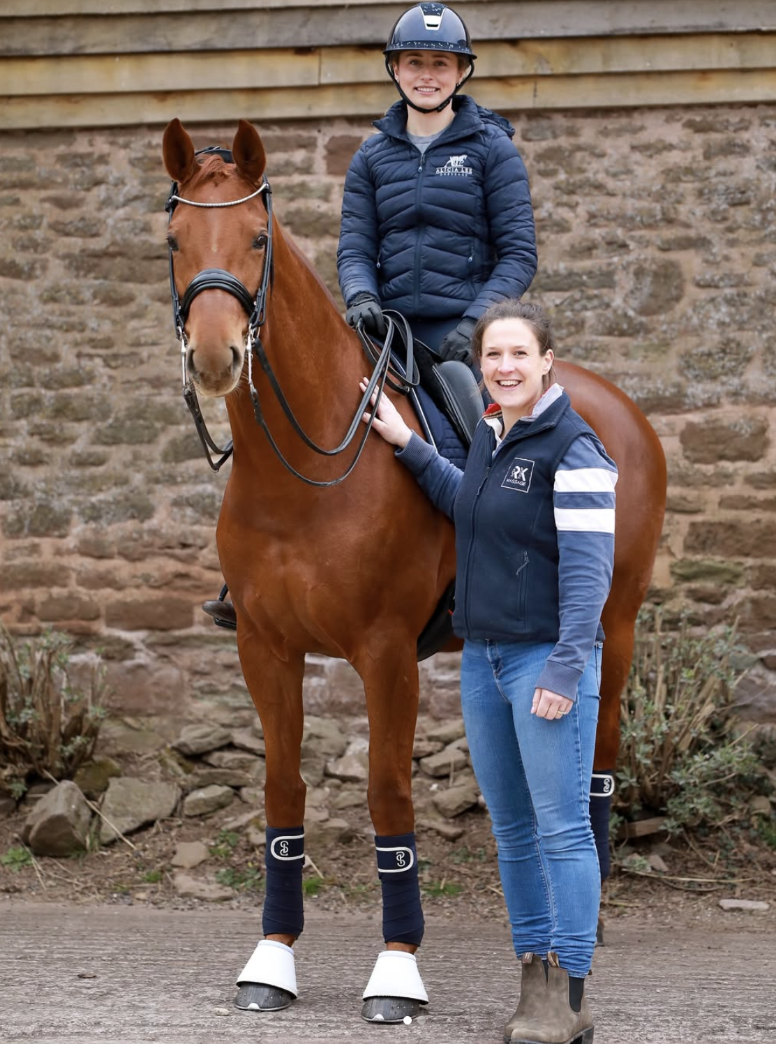 A young girl wearing a helmet and navy jacket riding a chestnut horse with white leg wraps, accompanied by a woman in a navy jacket and jeans standing next to the horse, outdoors with a brick wall background.