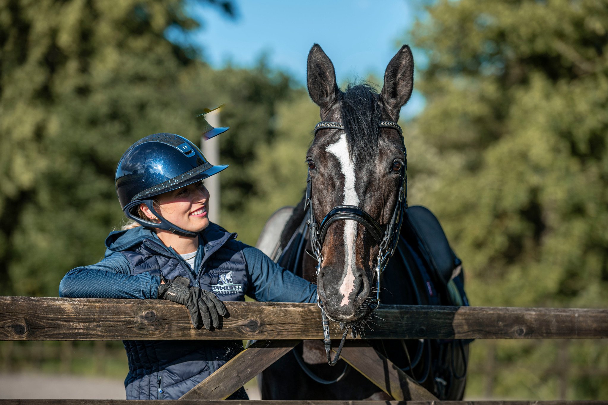 A woman wearing a helmet and riding gear smiling while leaning on a wooden fence next to a black and white horse.