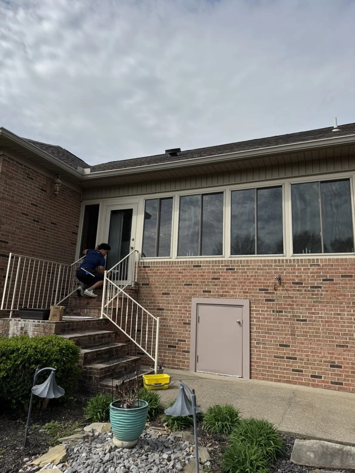A person is crouching on the stairs cleaning windows and door, outside a brick house with a large enclosed porch. There are plants and a garden area in the foreground, some gardening tools, and a cloudy sky above.