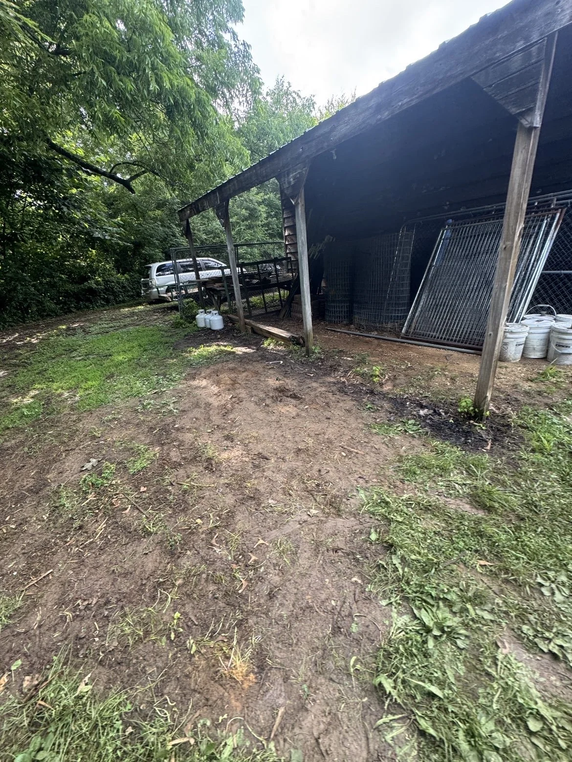 A rural outdoor scene featuring a weathered wooden shelter with metal fences inside, a dirt ground, and a vehicle parked underneath trees in the background. all previous debris is removed